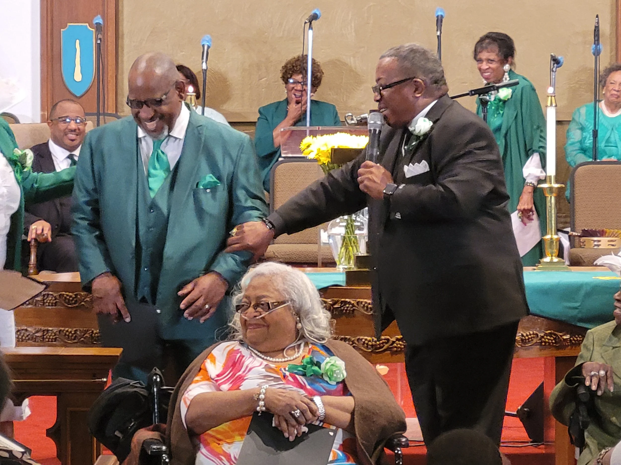 A man in a suit and glasses holds a microphone and taps the arm of another man in a teal suit, who is smiling, in a room with women in teal and green robes, chairs, microphones, and flowers.
