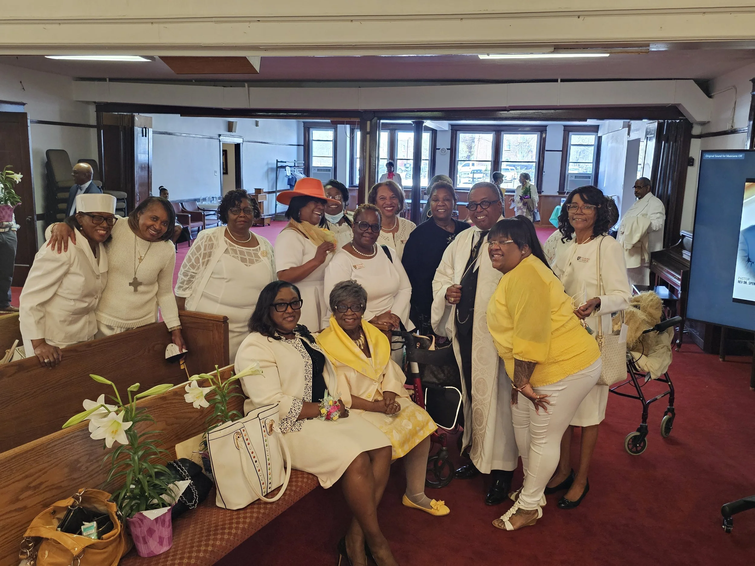 A group of 15 women and men gathered together indoors, smiling and posing for a photo, with some seated and others standing. Some women are dressed in white and yellow outfits, and one woman wears an orange hat. The background shows large windows and