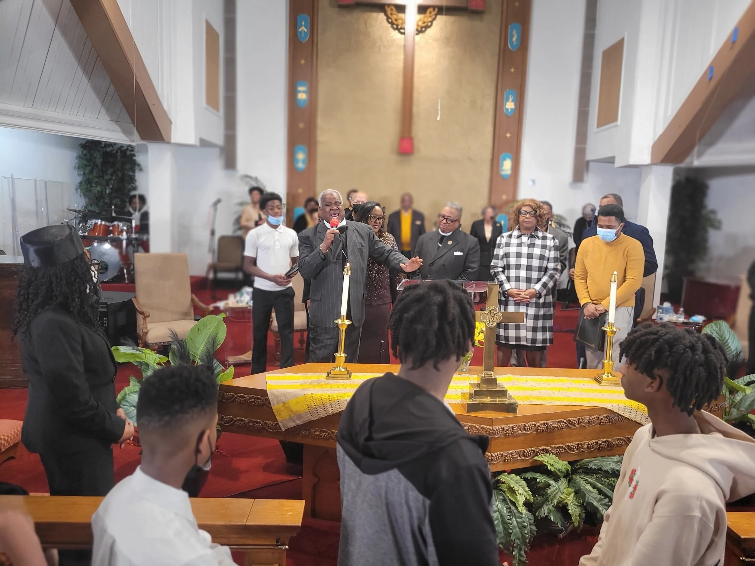A church service with a man speaking into a microphone at the altar, surrounded by choir members and congregation, with musical instruments in the background.