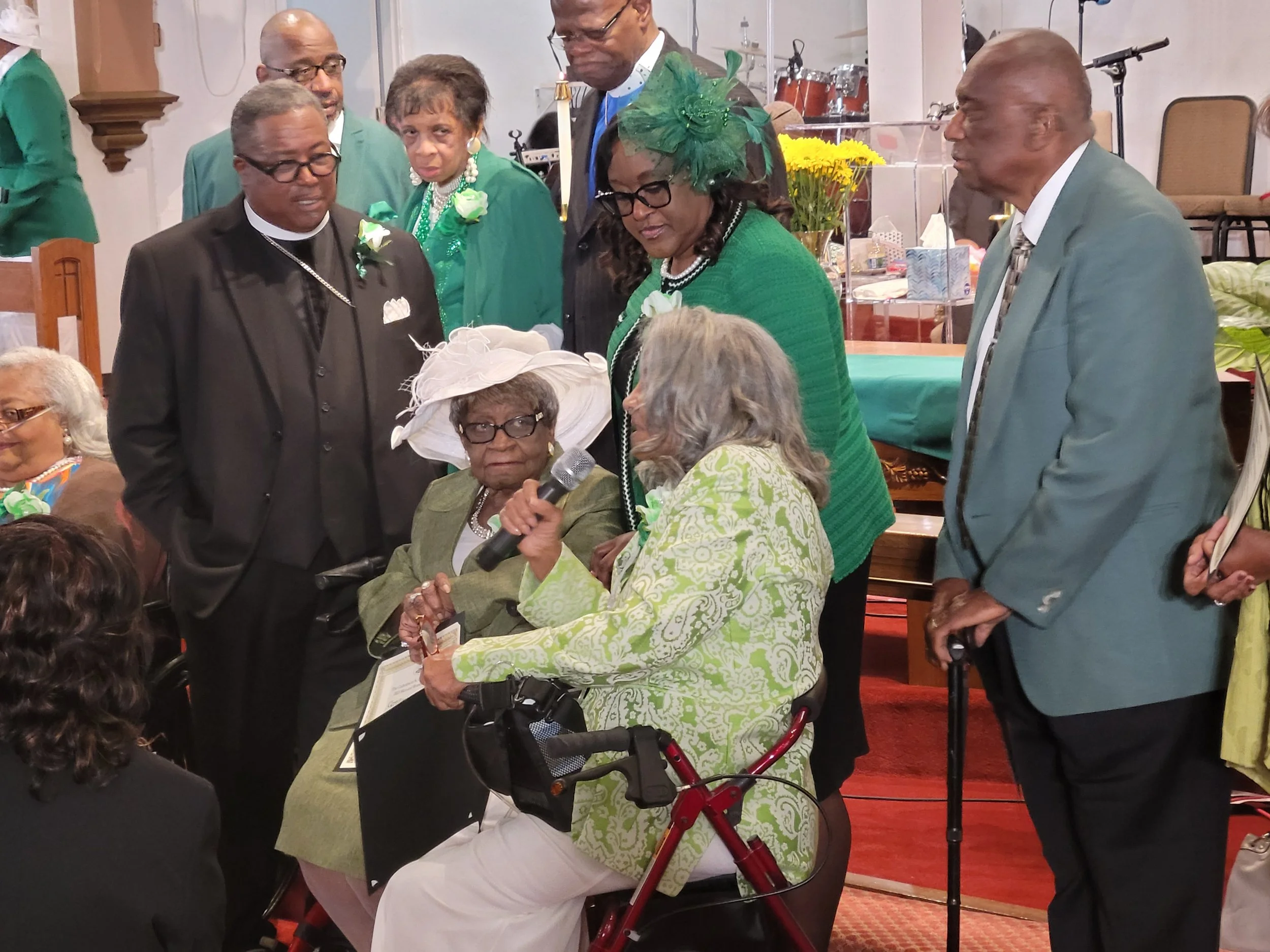 A group of elderly and middle-aged people at a gathering or celebration inside a church or community hall. A woman in a green outfit with glasses and a large green hat is speaking into a microphone held by another woman. The elderly woman is seated i