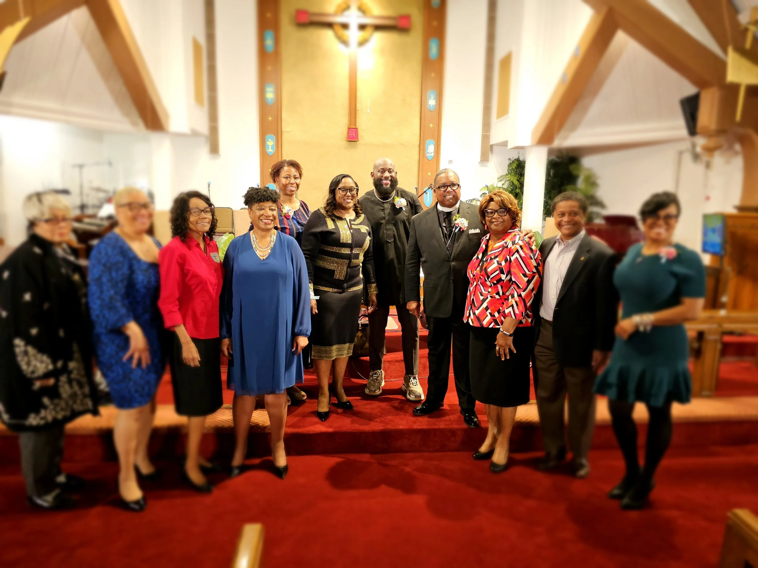 Group of people dressed in formal attire standing inside a church on red carpeted floor with a religious altar and cross behind them.