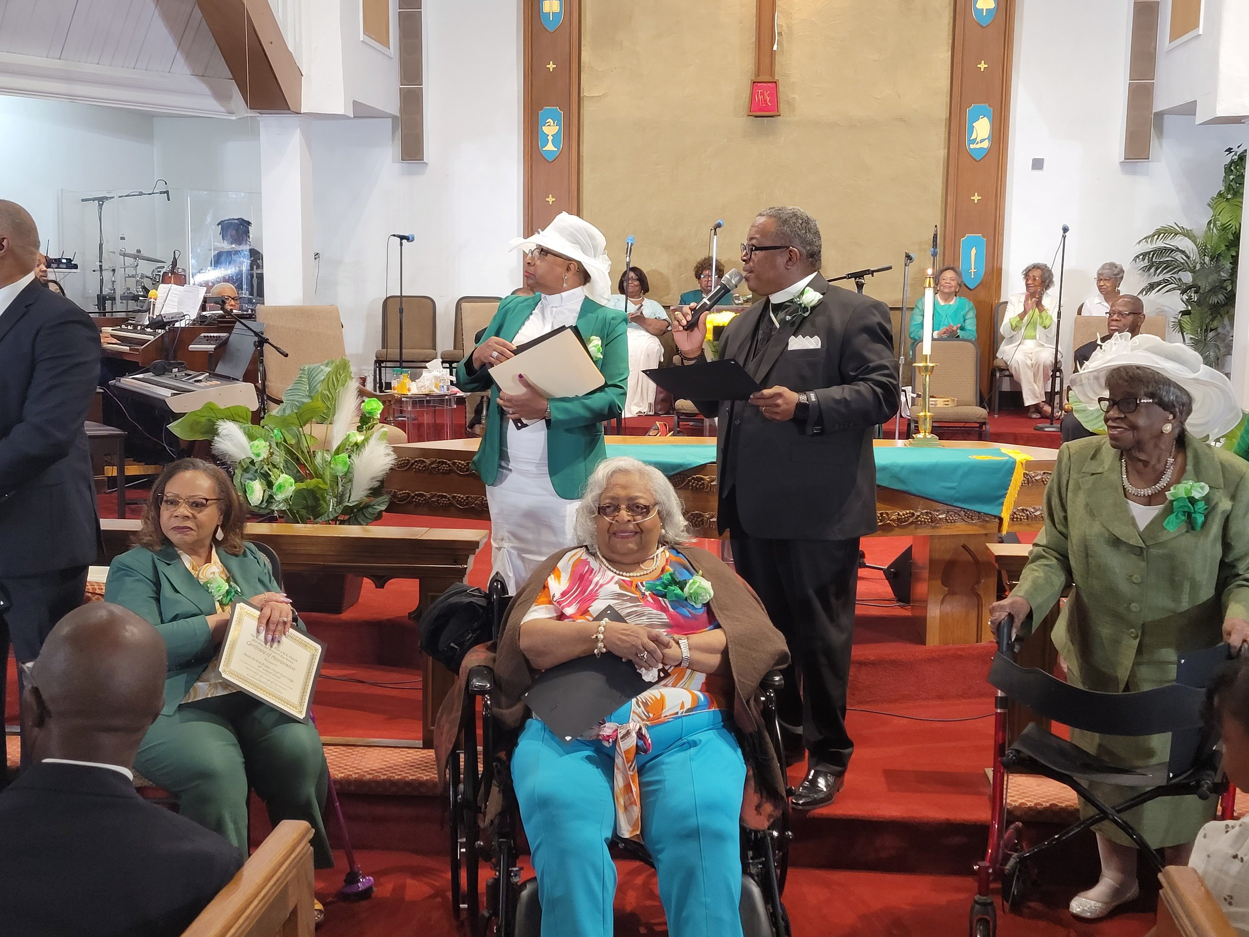 A diverse group of elderly people gathered in a church during a ceremony, some standing and others seated, with a man speaking into a microphone and women dressed in colorful outfits and hats.