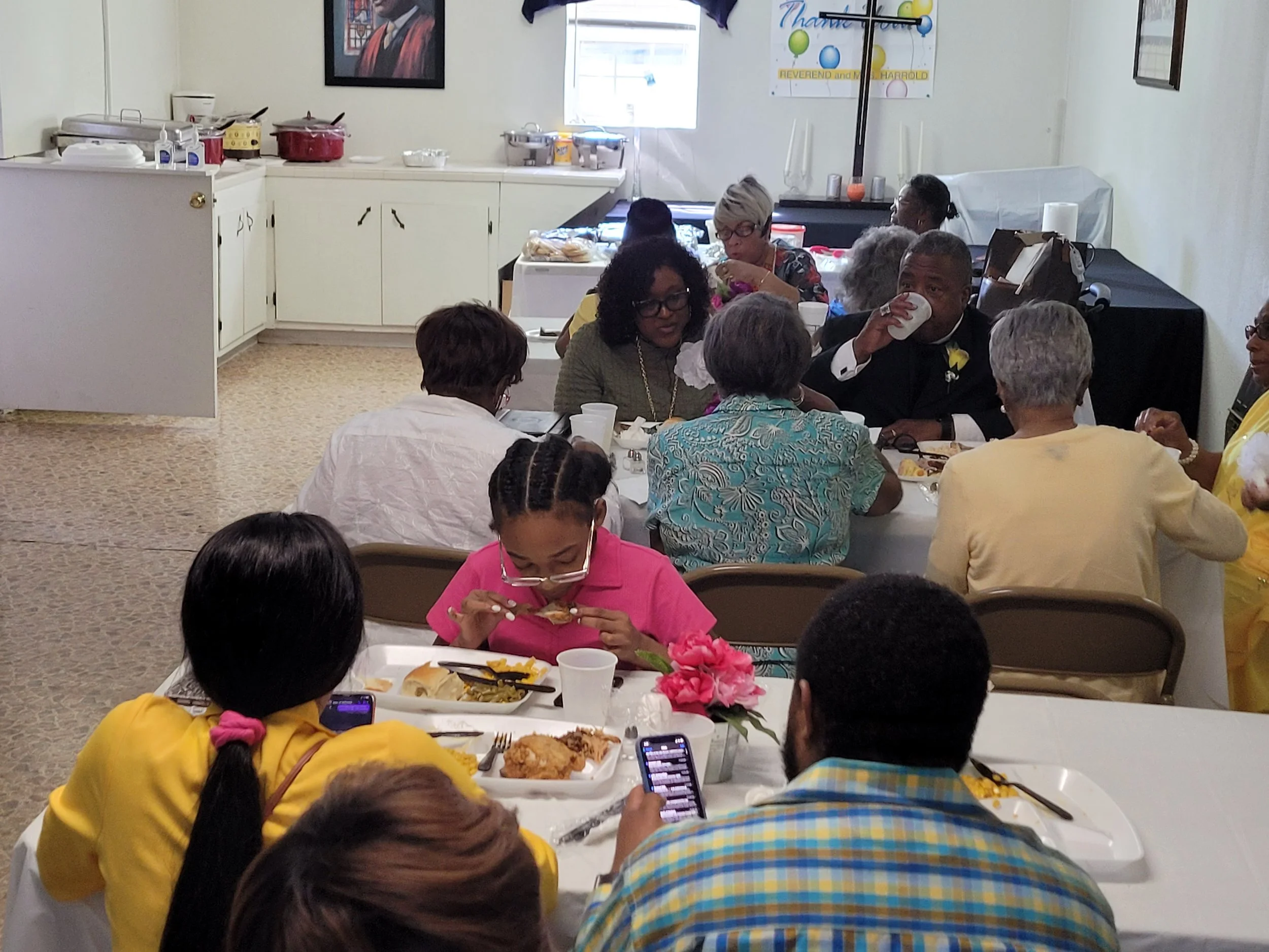 People sitting at a table during a gathering, eating food, with some using phones, in a decorated room, possibly for a celebration.