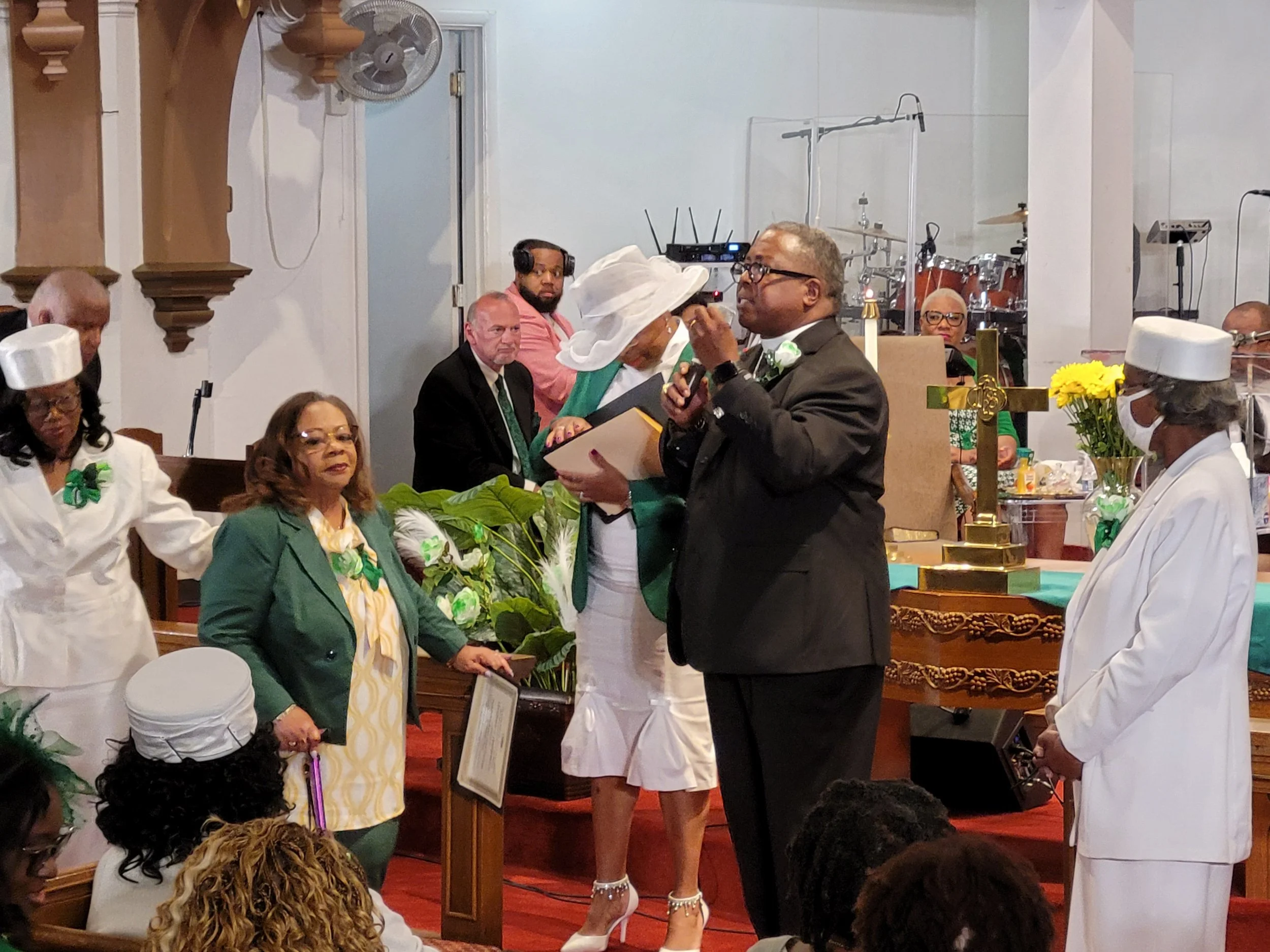 A church service with several people dressed in formal and religious attire, including a man in a suit speaking into a microphone, with others standing around him, some wearing white robes and hats, and a cross on the altar in the background.