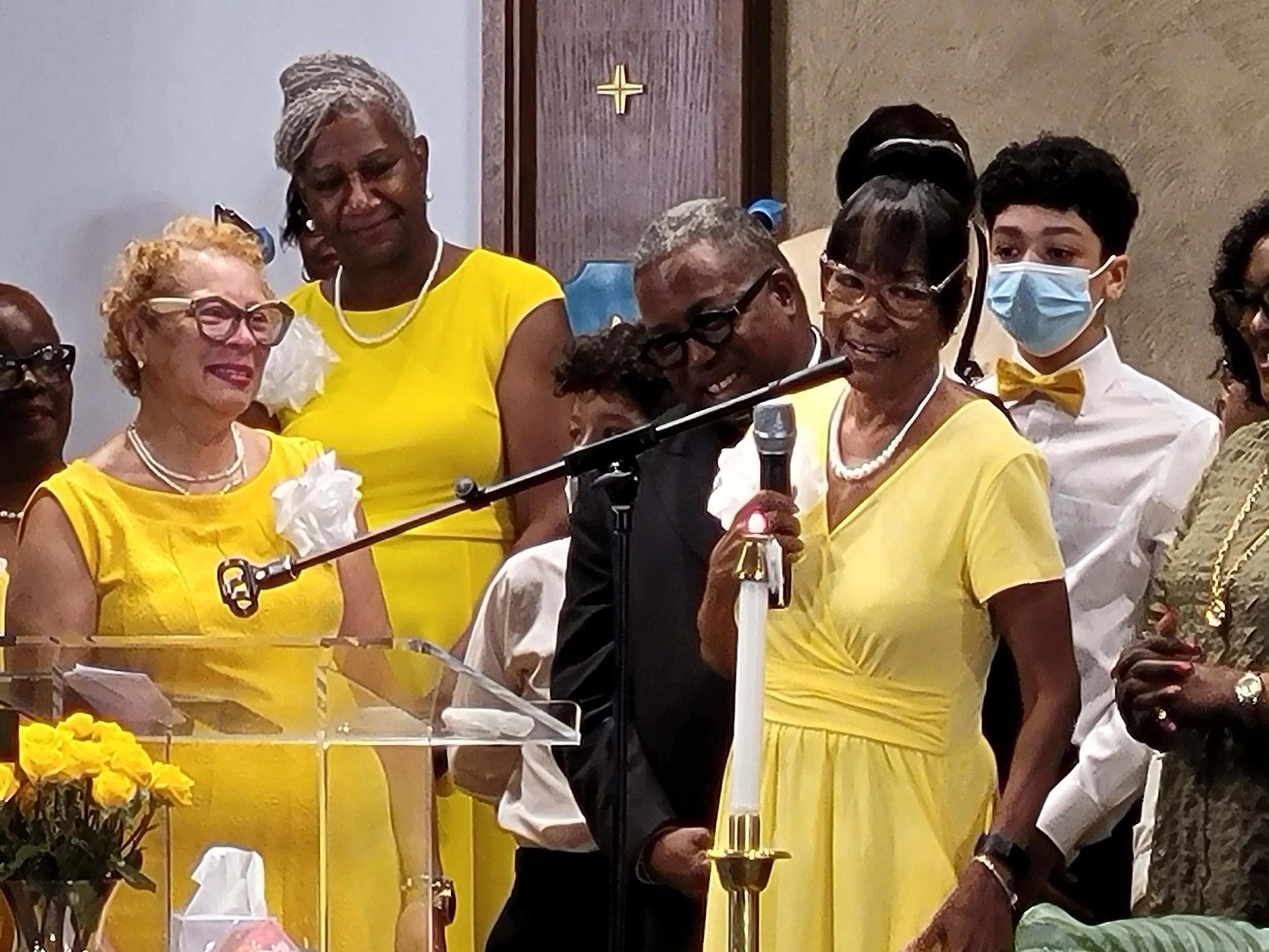 A group of people at a formal event, with most wearing yellow dresses or shirts. One woman is speaking into a microphone, holding it with both hands, while others are smiling and listening. A man in a black suit and glasses is standing nearby. Some i