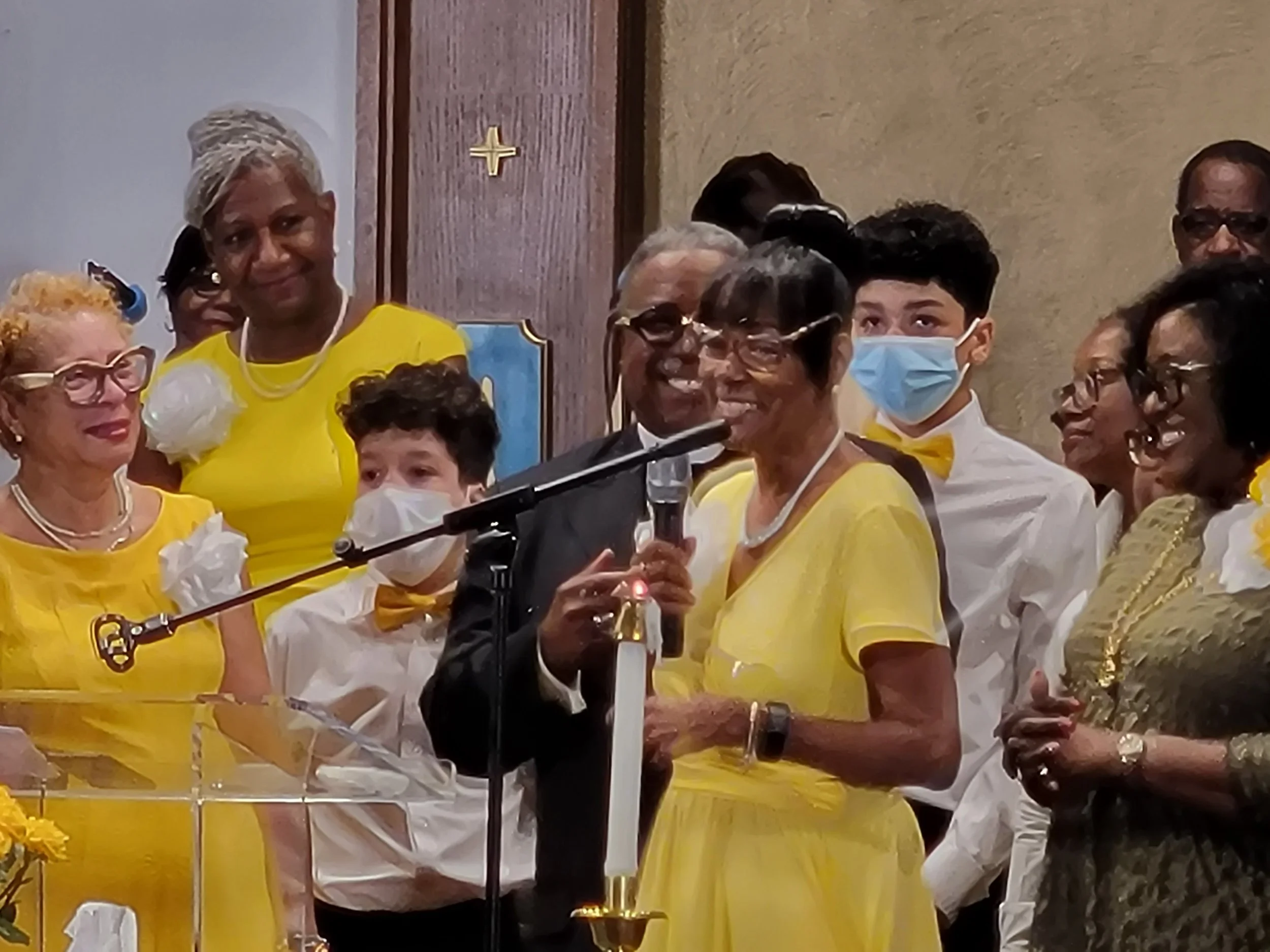 A group of people, all dressed in yellow or gold, gathered at a church event. A woman is speaking at a microphone, and some individuals are wearing face masks.