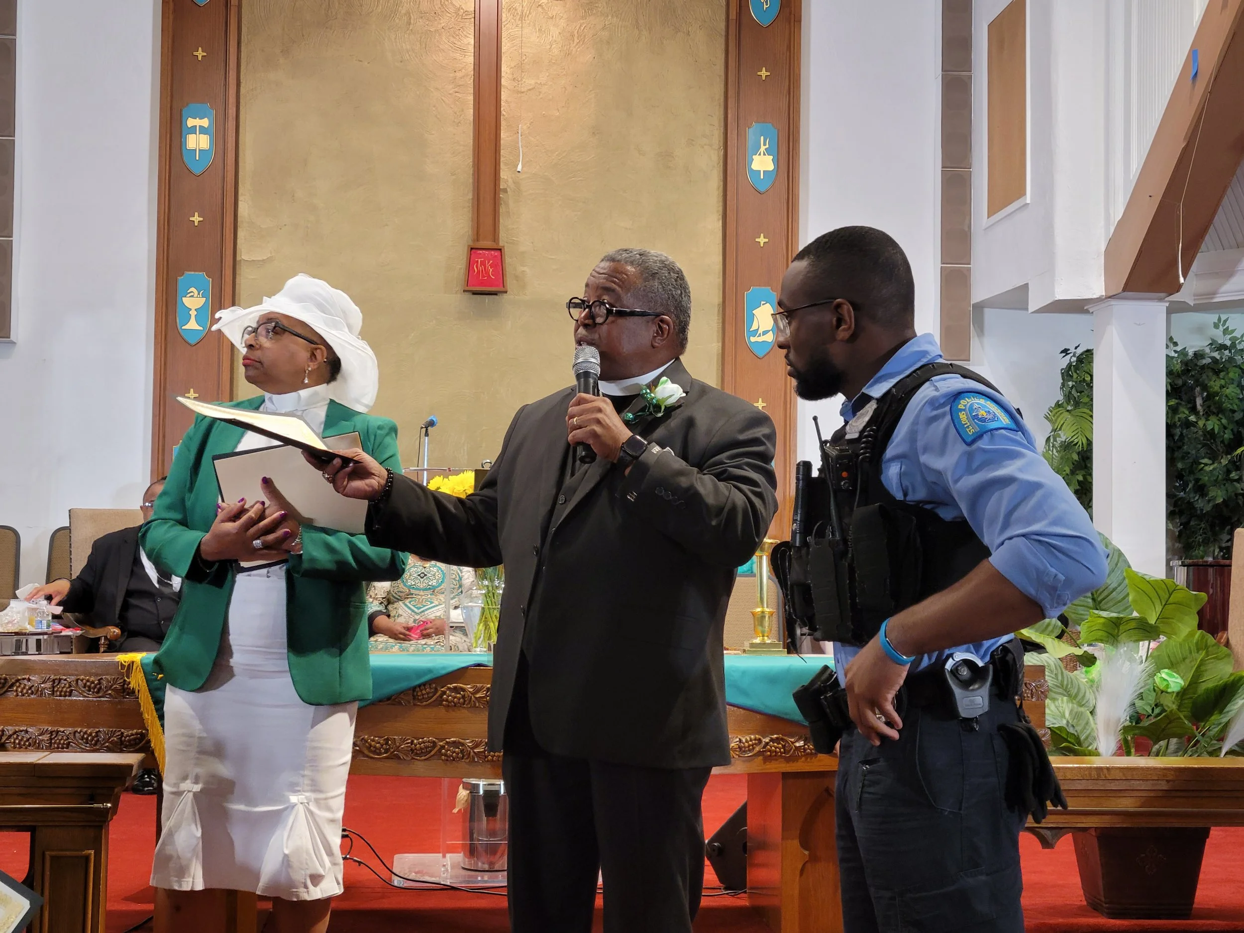 A man in a black suit with glasses and a white flower pinned to his lapel is speaking into a microphone while holding a document, standing at a church ceremony. A woman in a white hat, green blazer, and white skirt stands nearby, holding a folder, an