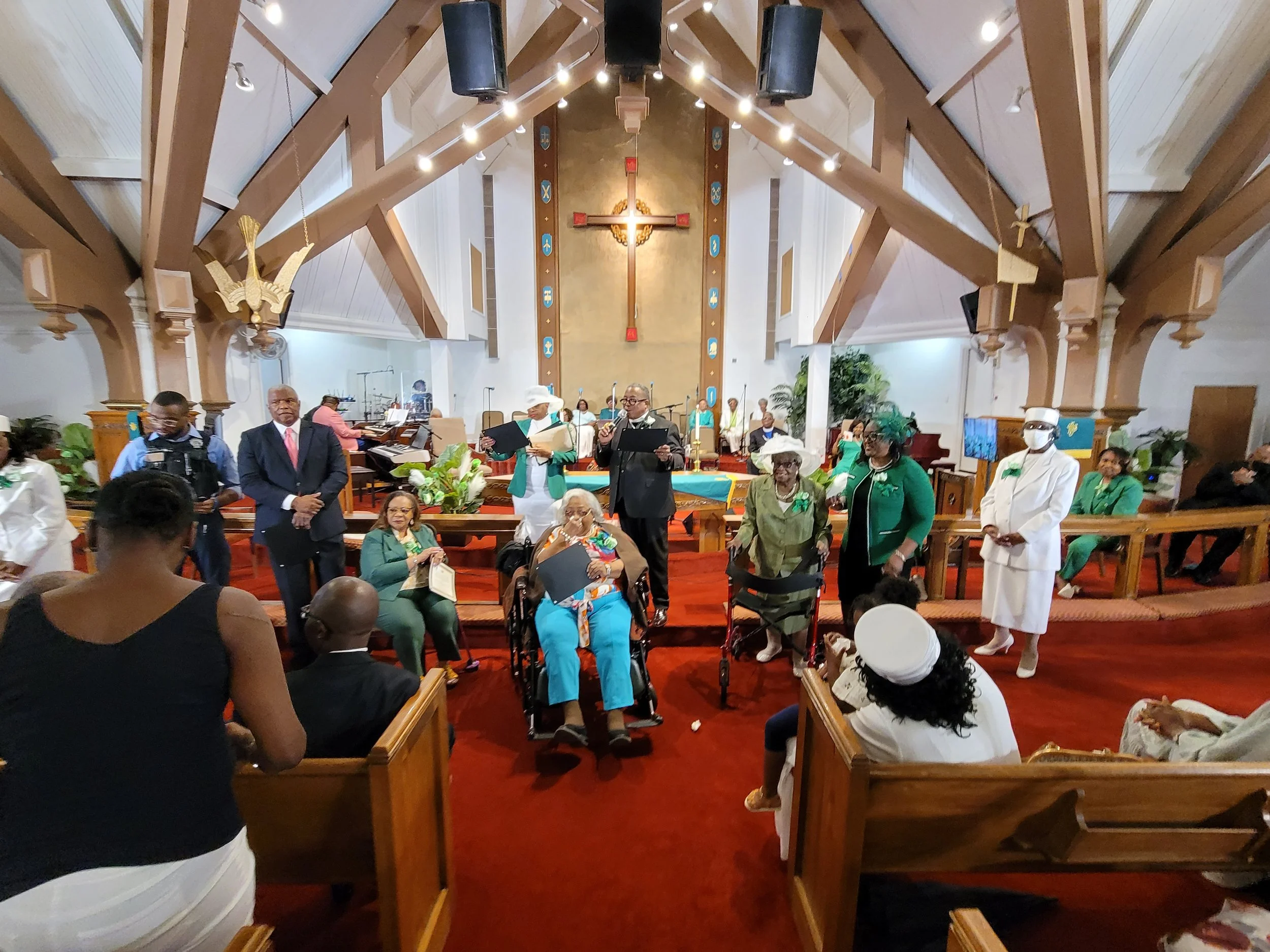 People gathered inside a church, some standing and others seated, with a large cross and religious symbols on the wall behind the altar. The church has wooden beams and a red carpet floor.