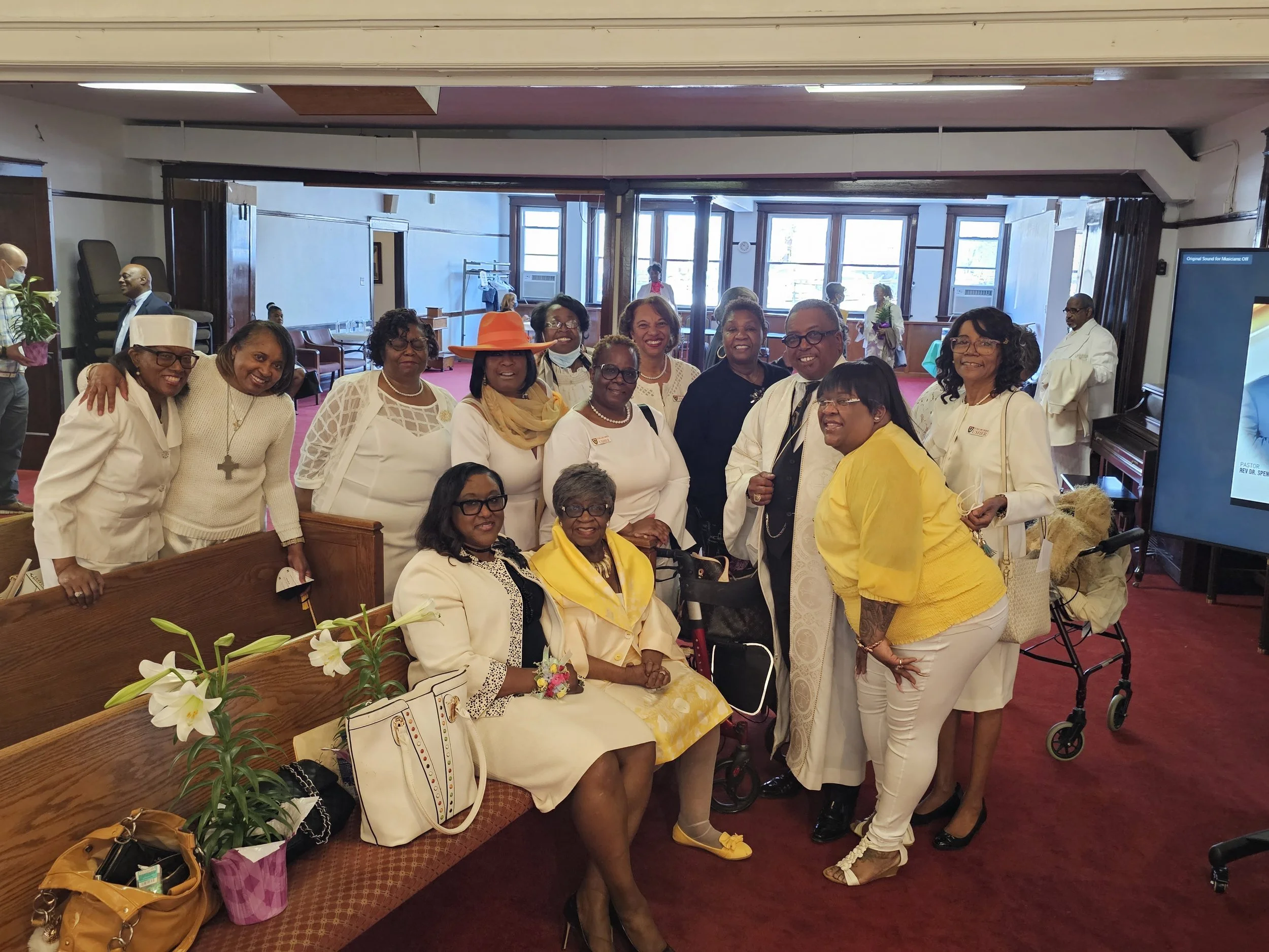 Group of mostly women dressed in formal white, yellow, and black attire, posing together inside a church or community hall for a photo, with some seated and some standing, and a few with leis or accessories.