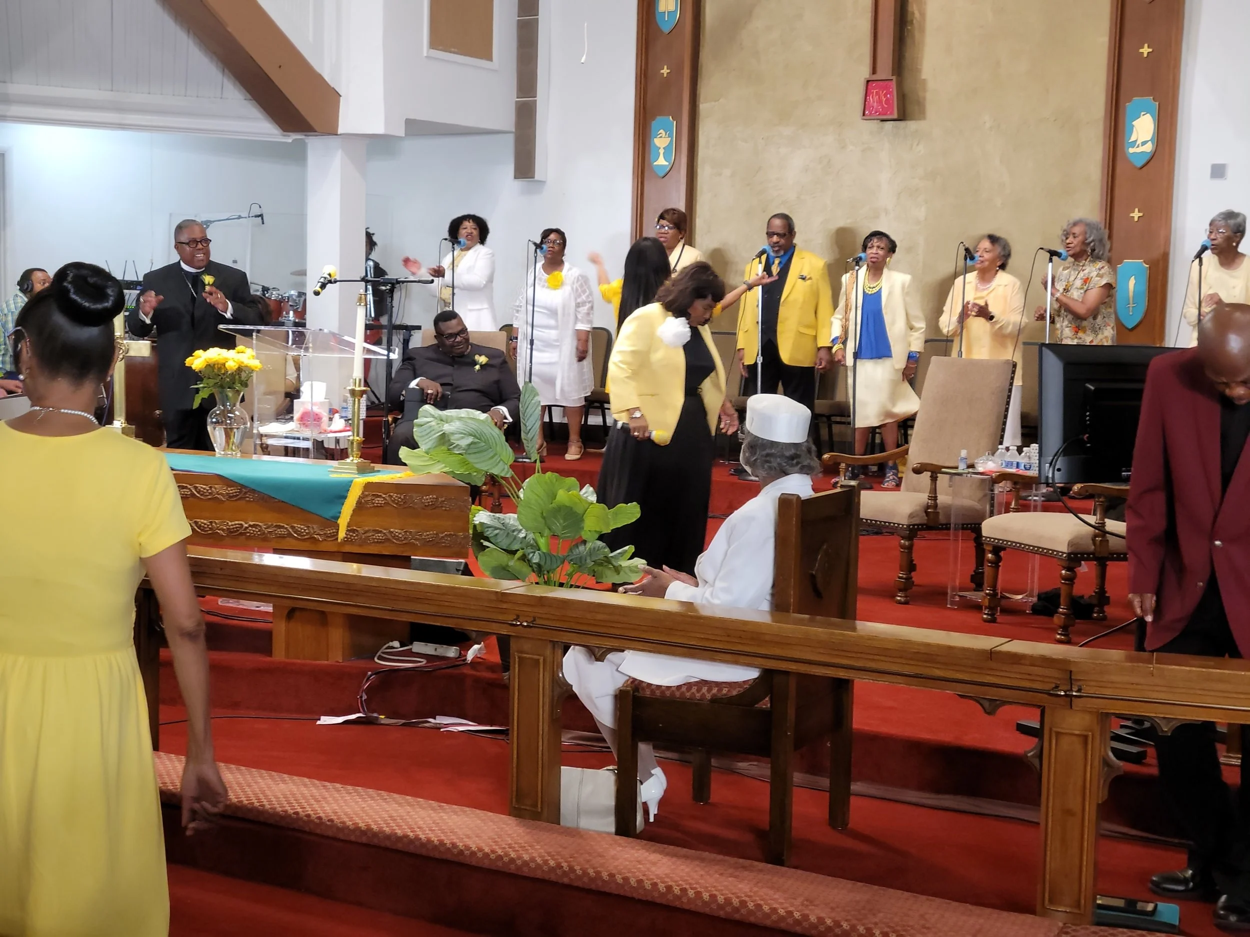 Church choir and congregation during service, with choir members singing on stage, and audience members in yellow and white attire.