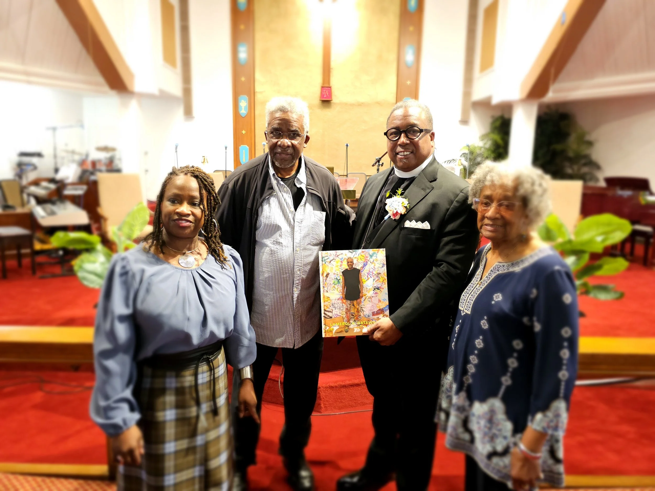 Four people standing inside a church with a man in a suit holding a colorful book. The church has a red carpet, wooden pews, and religious symbols on the wall.