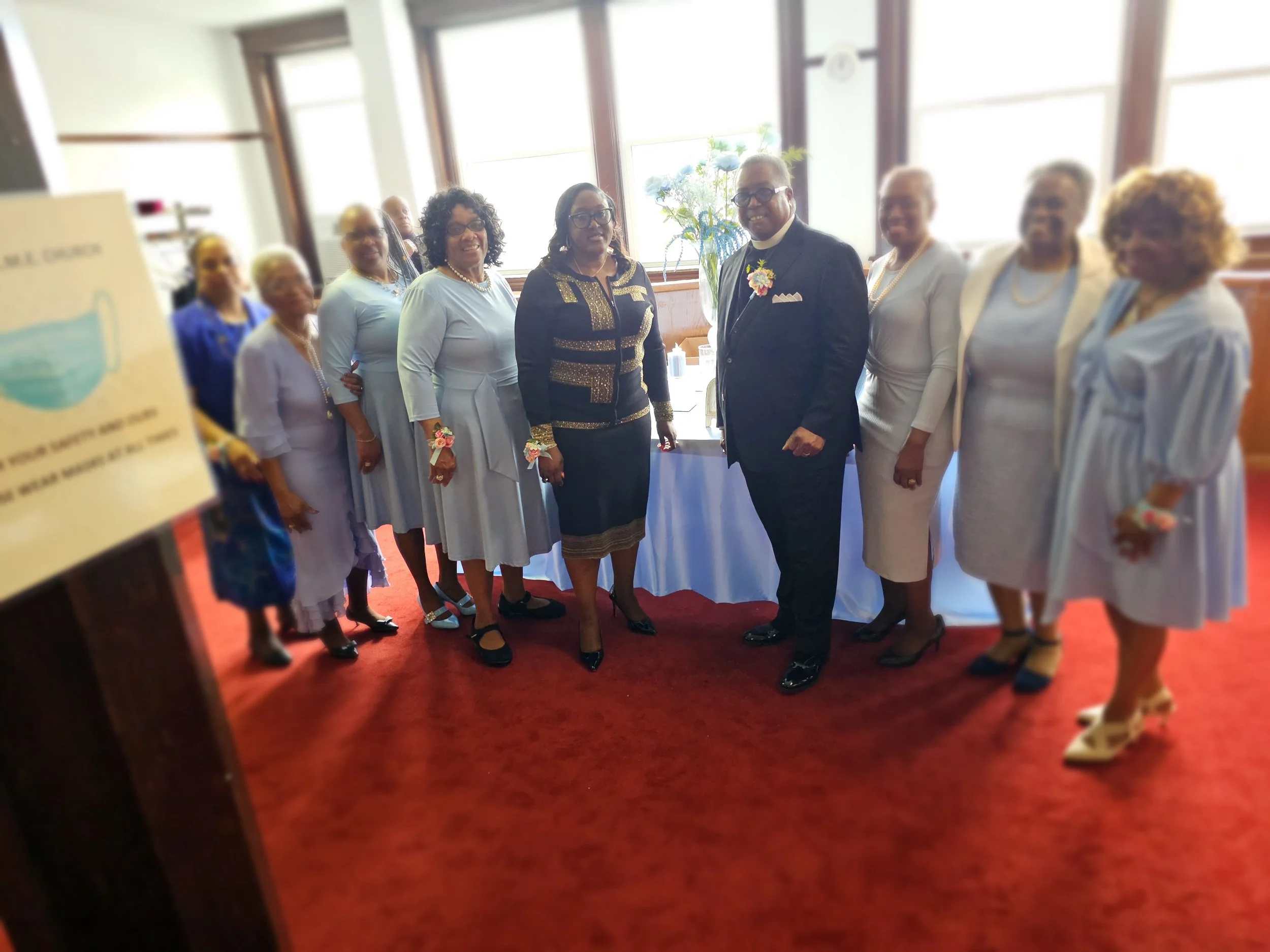 Group of men and women dressed in formal attire standing together in a room with a red carpet, large windows, and a table with a floral arrangement in the background.