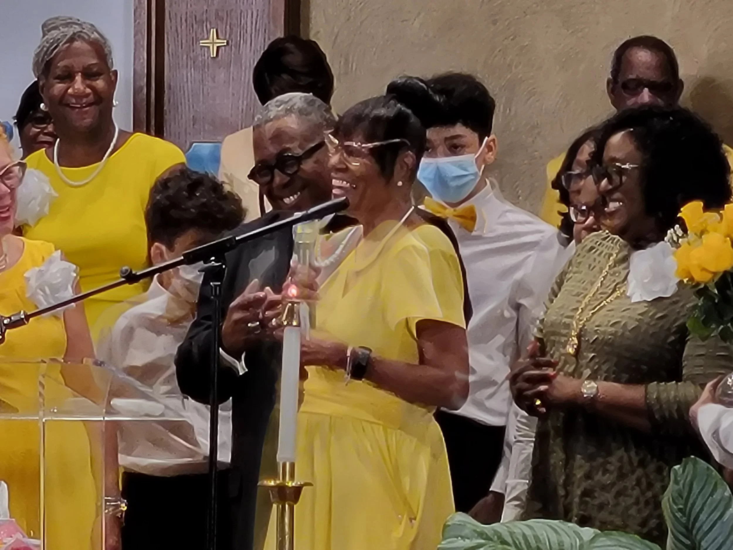 Group of people celebrating, with some holding glasses, dressed in yellow and white, one person wearing a face mask, at a gathering with smiling faces.