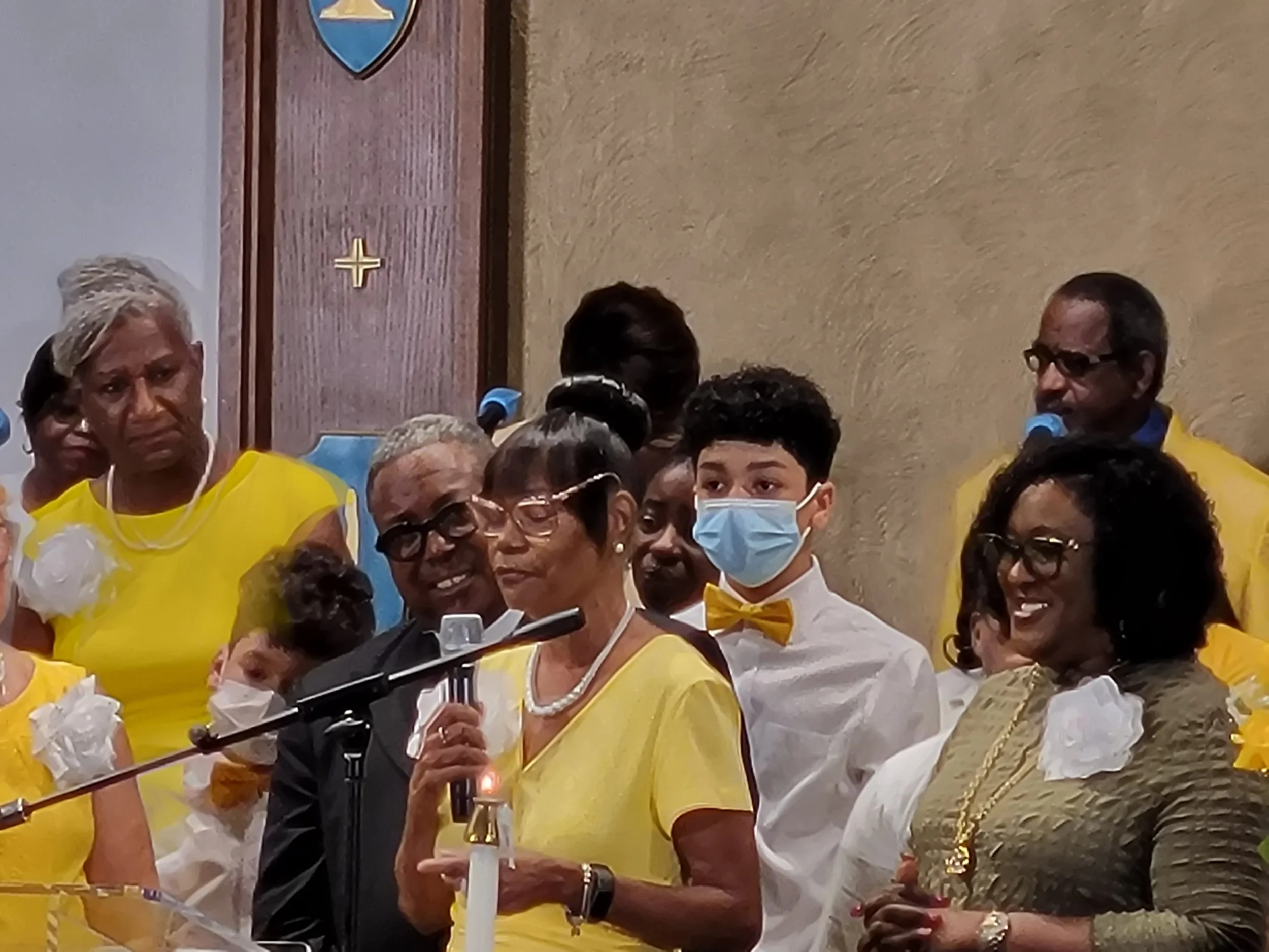 Group of people standing on a stage during a ceremony, some wearing yellow outfits, one woman speaking into a microphone, some wearing face masks, in an indoor setting.