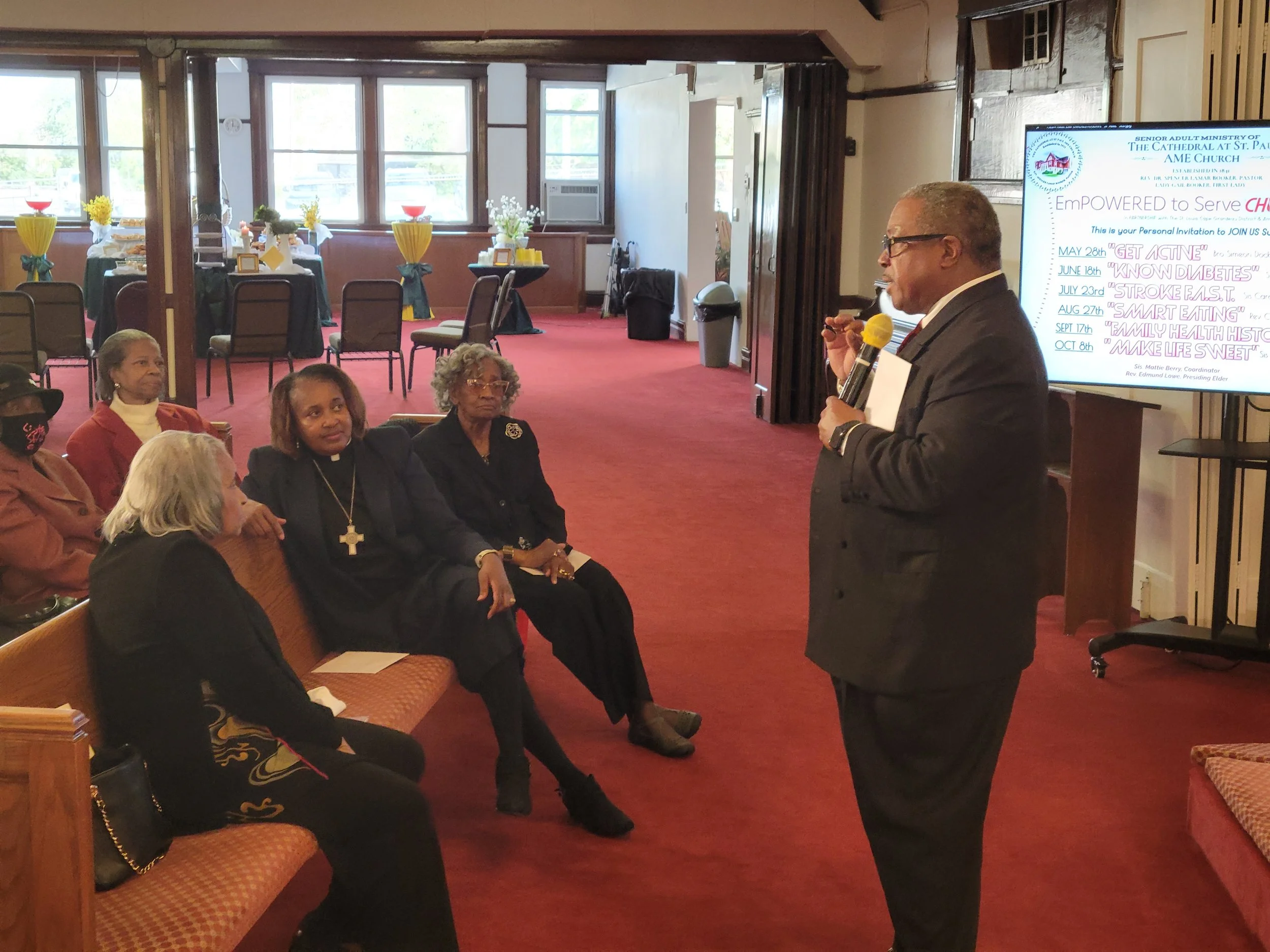 A man in a suit holds a microphone and speaks to a seated audience in a room decorated with tables and chairs. A large screen displays a presentation slide with event details and a schedule, including topics like diabetes, stroke, and family health.