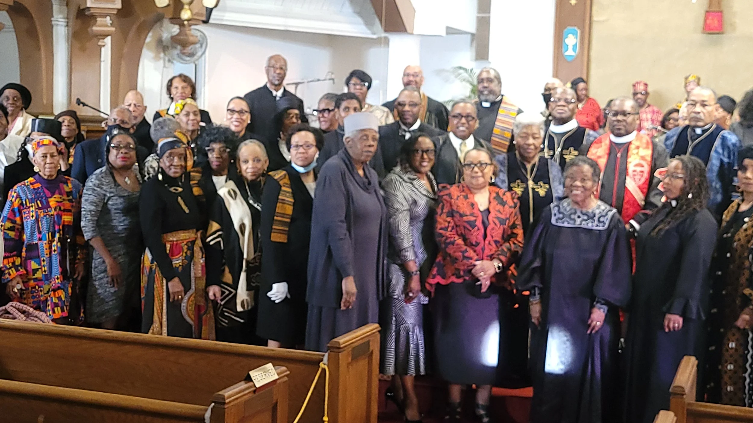 A large group of diverse people, dressed formally and wearing traditional African garments, gathered inside a church for a celebration or special event.