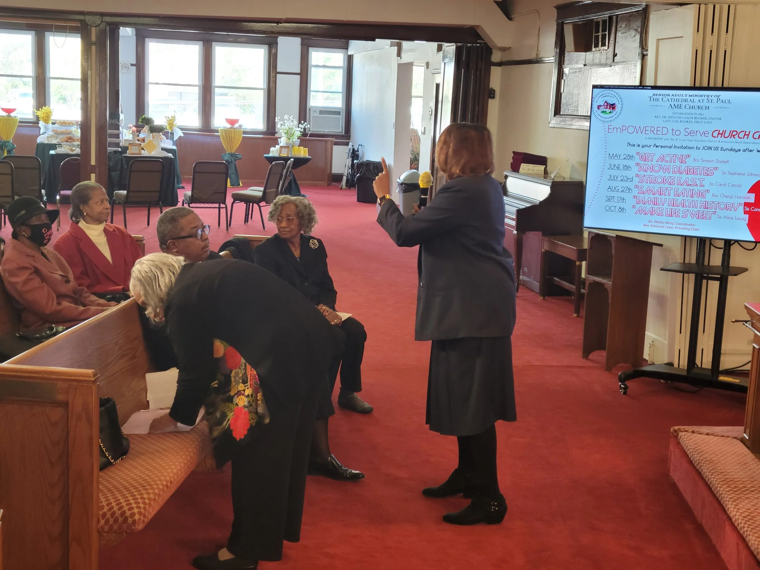 A woman in a dark blazer and long skirt speaking with a microphone to a group of women seated on a church pew inside a church. The background has a screen displaying event information and decorated tables with balloons and flowers.