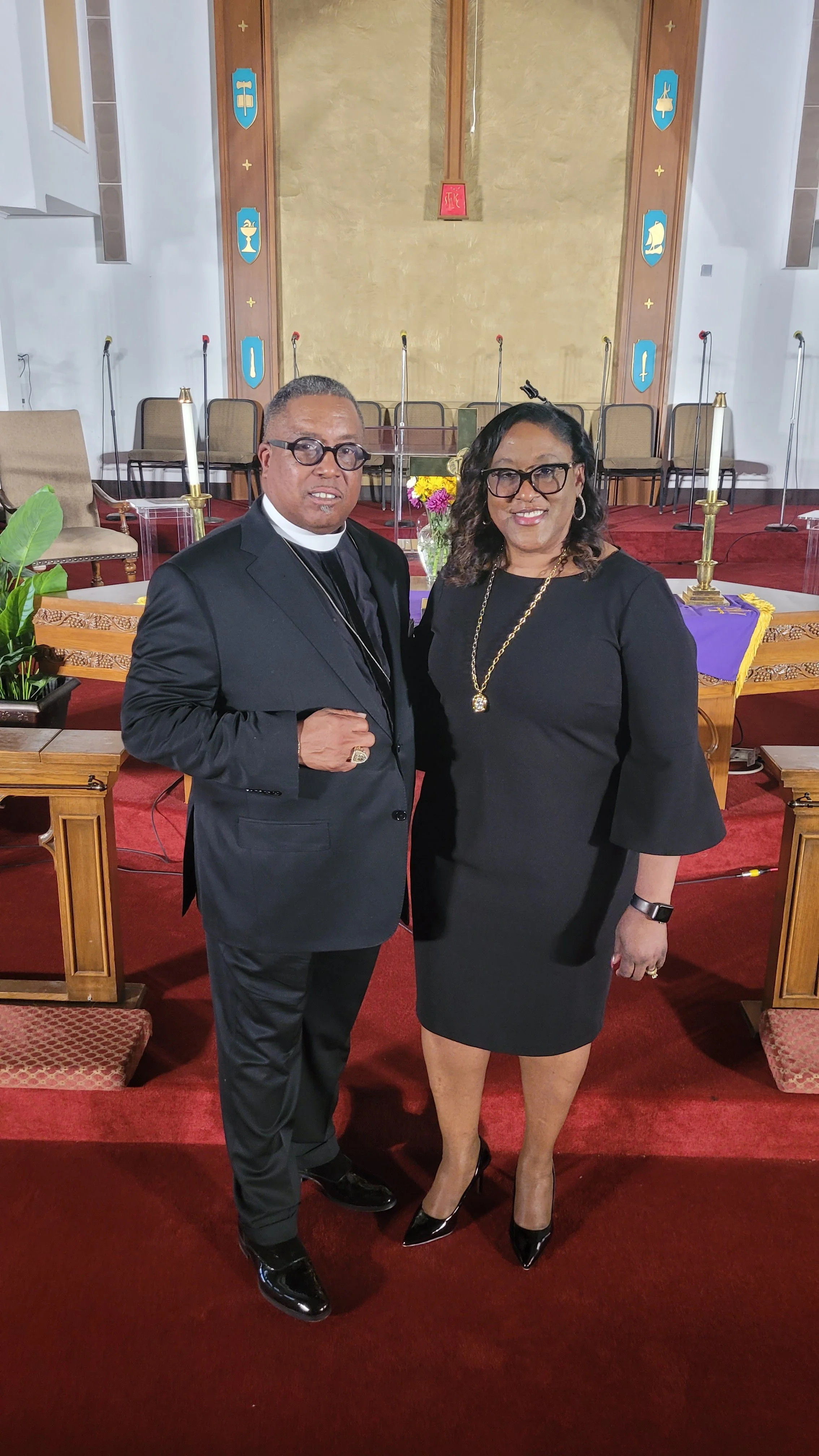 A man and woman standing together inside a church. The man is dressed in a black suit with clerical collar and glasses. The woman is wearing a black dress, heels, and glasses. They are smiling and posing for the photo in front of a stage with candles