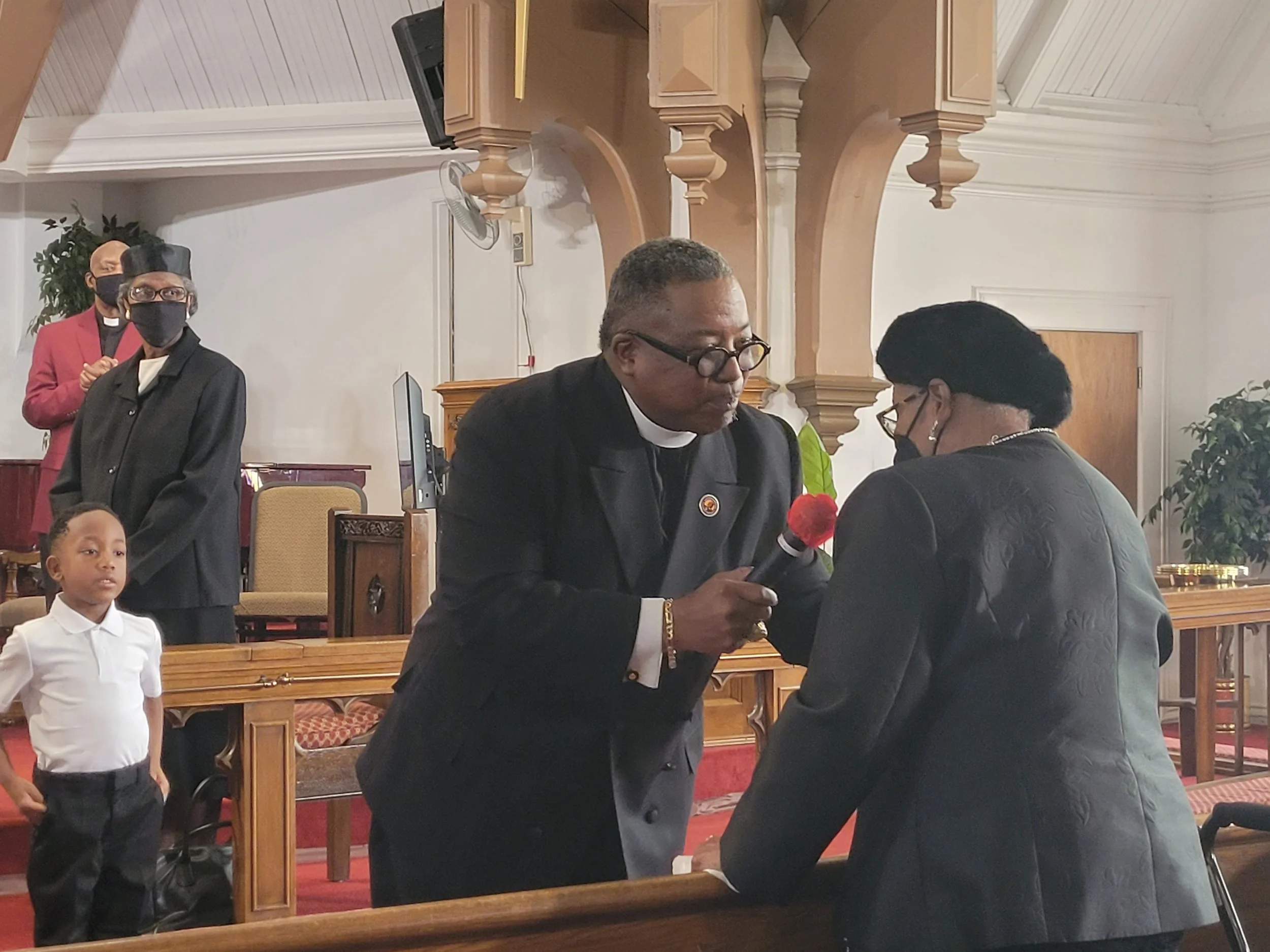 A man in clergy attire is speaking into a microphone held by a woman in a black jacket within a church setting, with children and other adults observing.