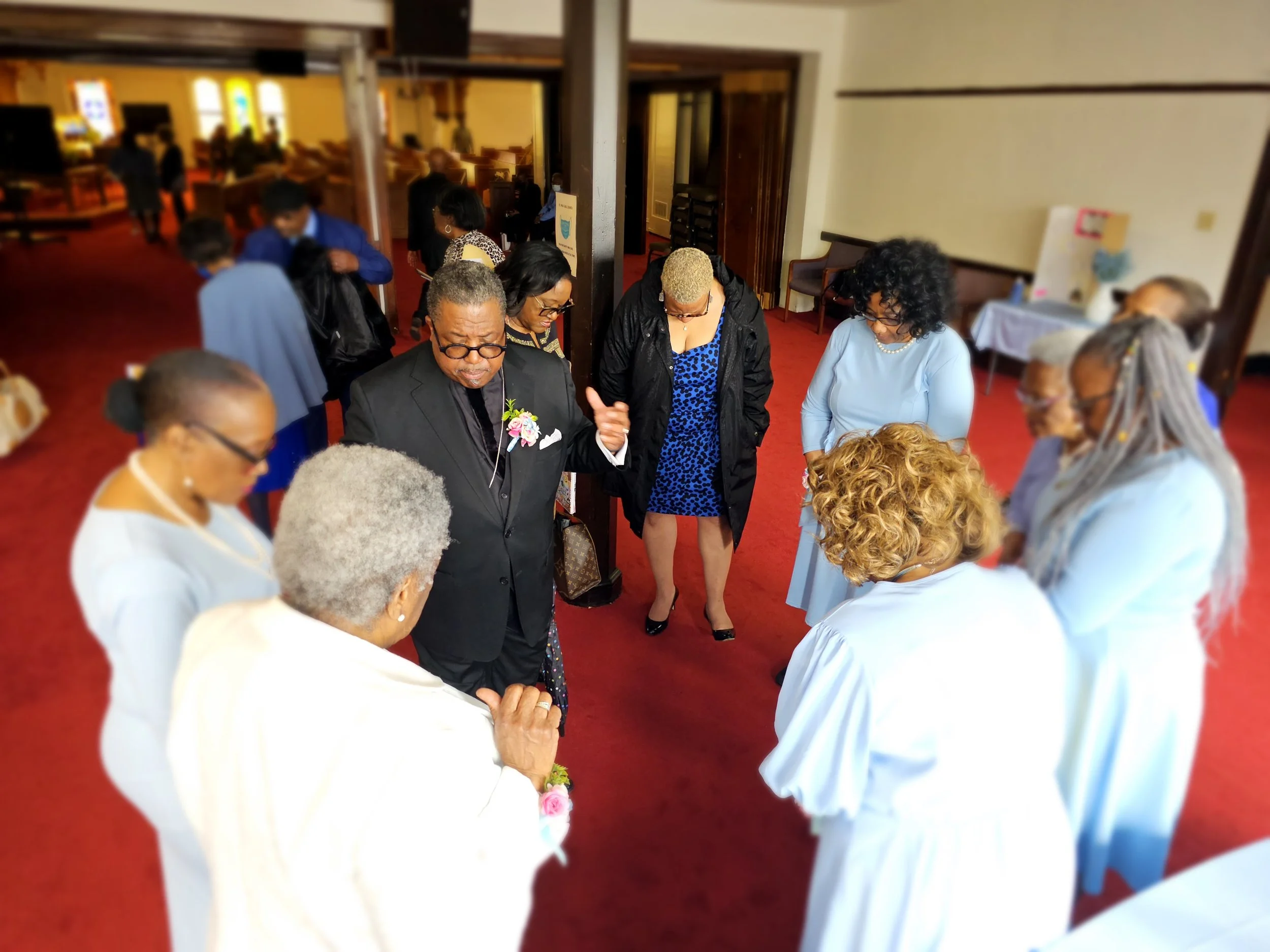 Group of people praying together in a church, with some dressed in white robes and others in formal attire, in a circle on a red carpet.
