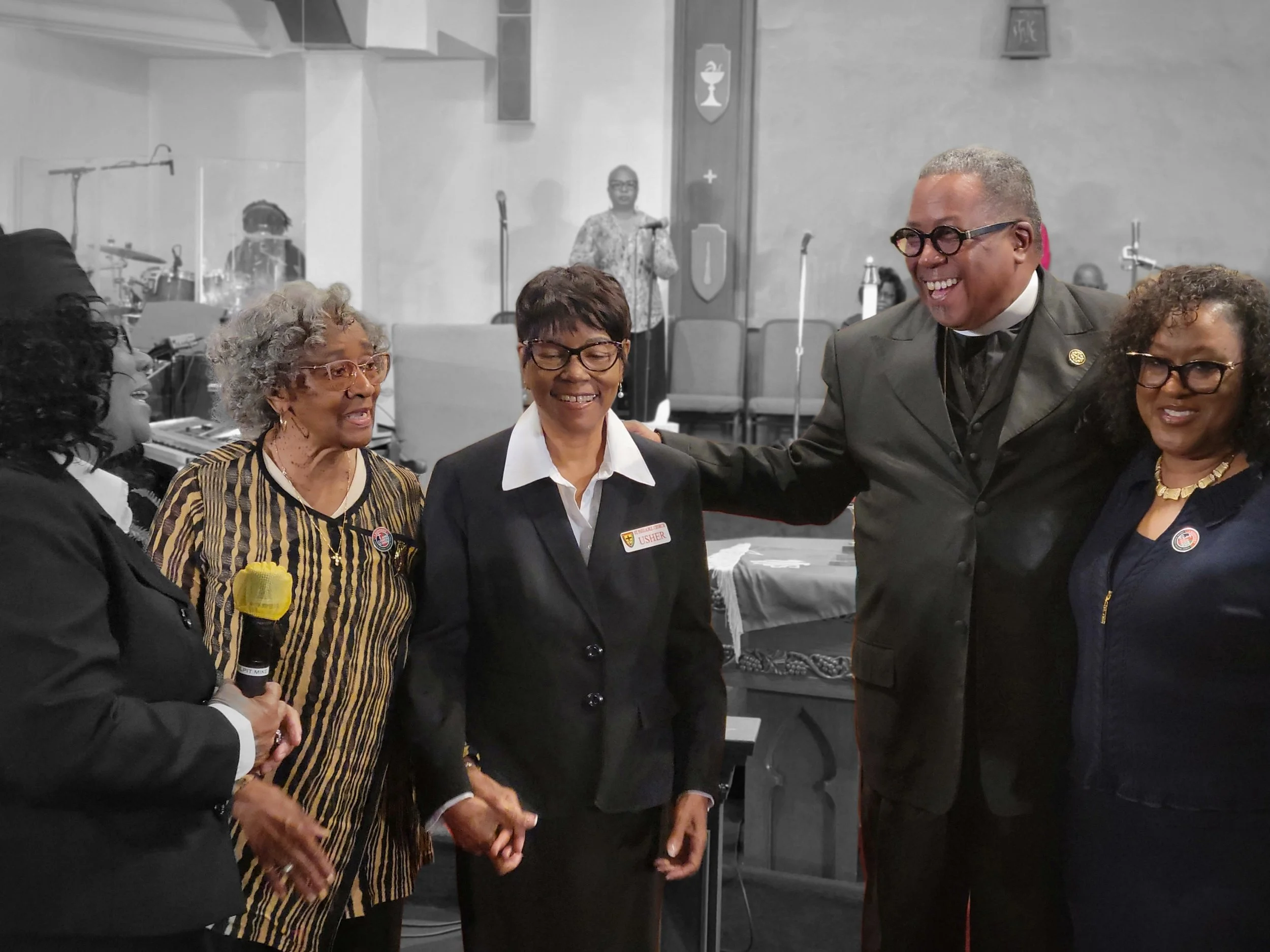 Group of five people, four women and one man, smiling and engaging in conversation at a formal event. The woman second from the right is wearing a black blazer with a badge, and the man is in a black suit with clerical collar. In the background, ther