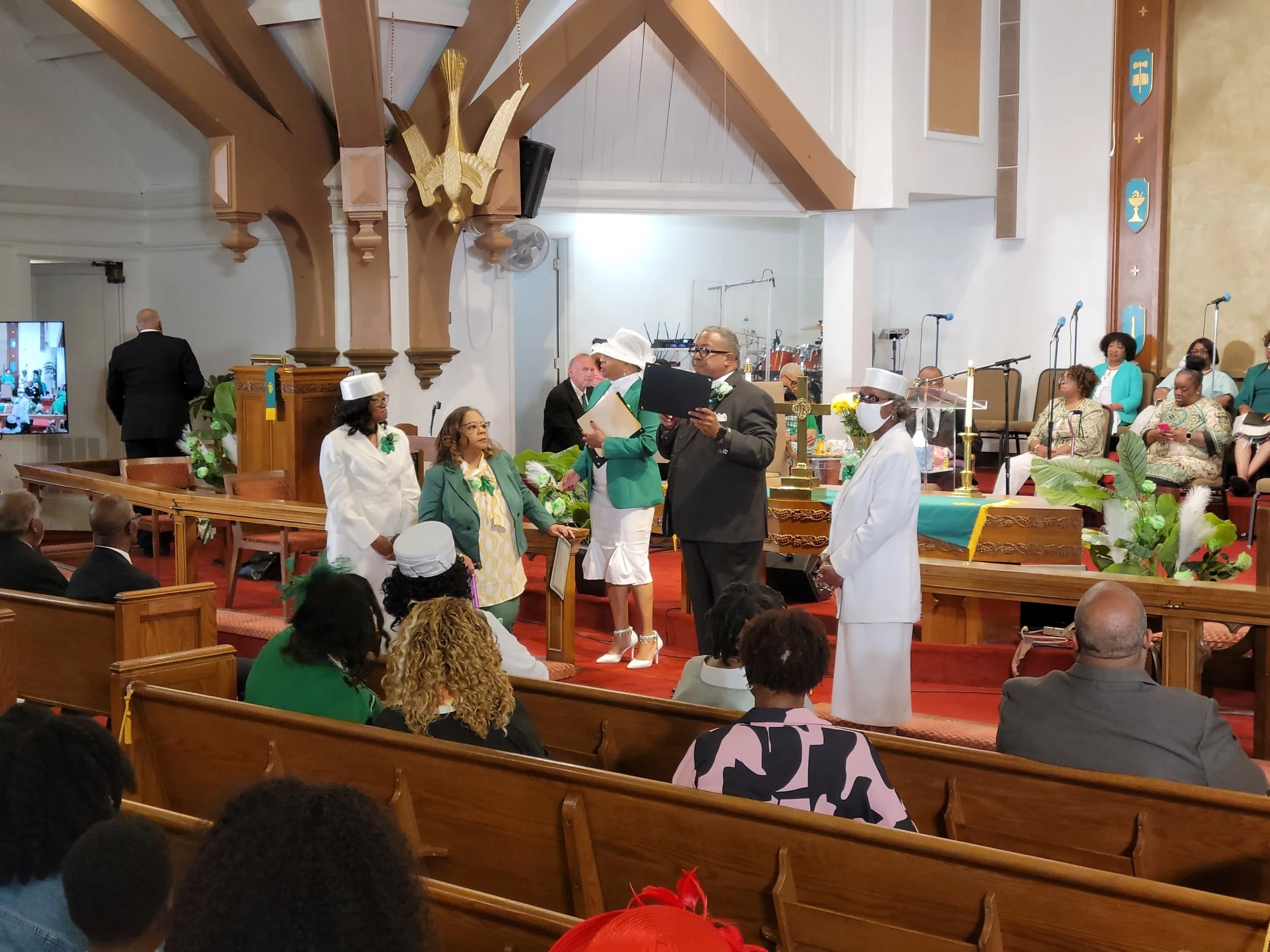 A church ceremony with several people standing in front of the altar, some dressed in white and green, and others in dark suits. There are people seated in the pews watching the event, and a choir seated in the background. The church has wooden pews,