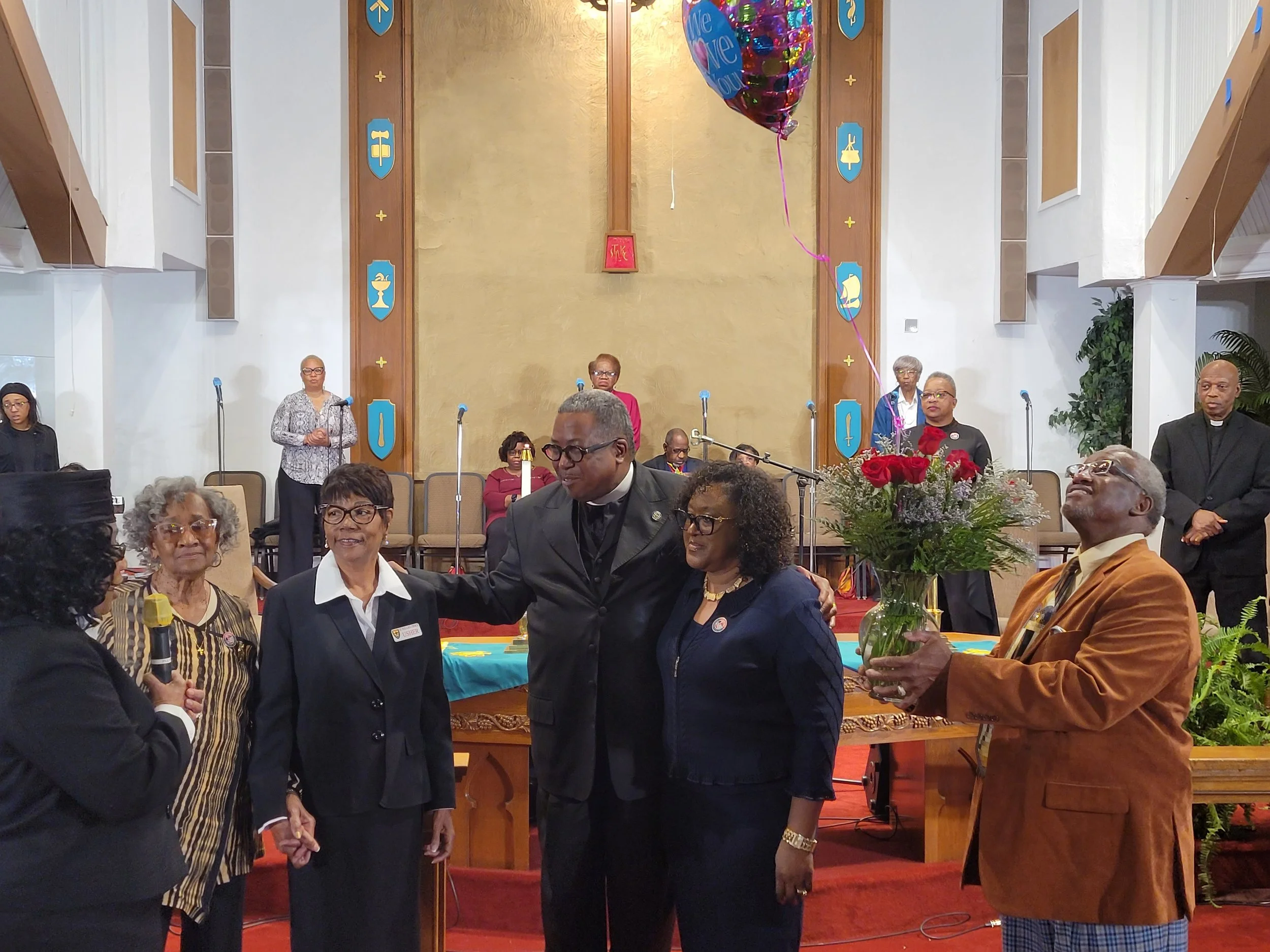 People gathered in a church, some standing on stage and some in the audience. A man with glasses and a brown jacket holds a bouquet of flowers. A woman in a dark blazer and glasses talks into a microphone, and another woman in a dark blazer with glas