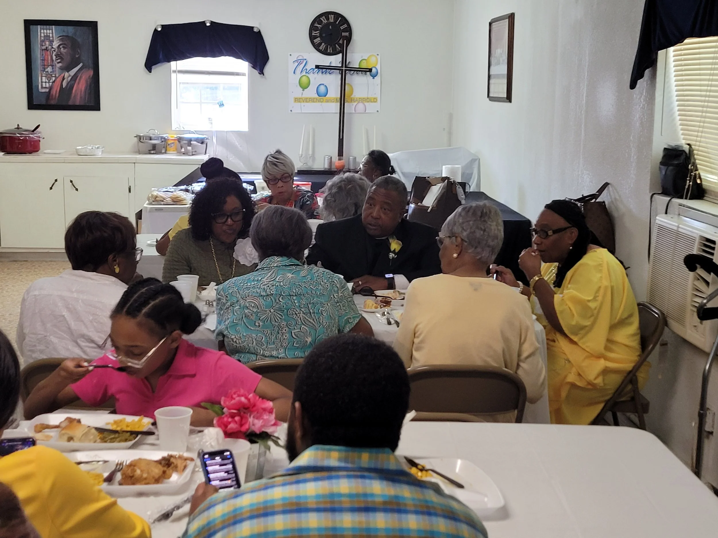 A group of people gathered at a social event, sitting around tables with food, in a room with artwork on the walls and an air conditioning unit.