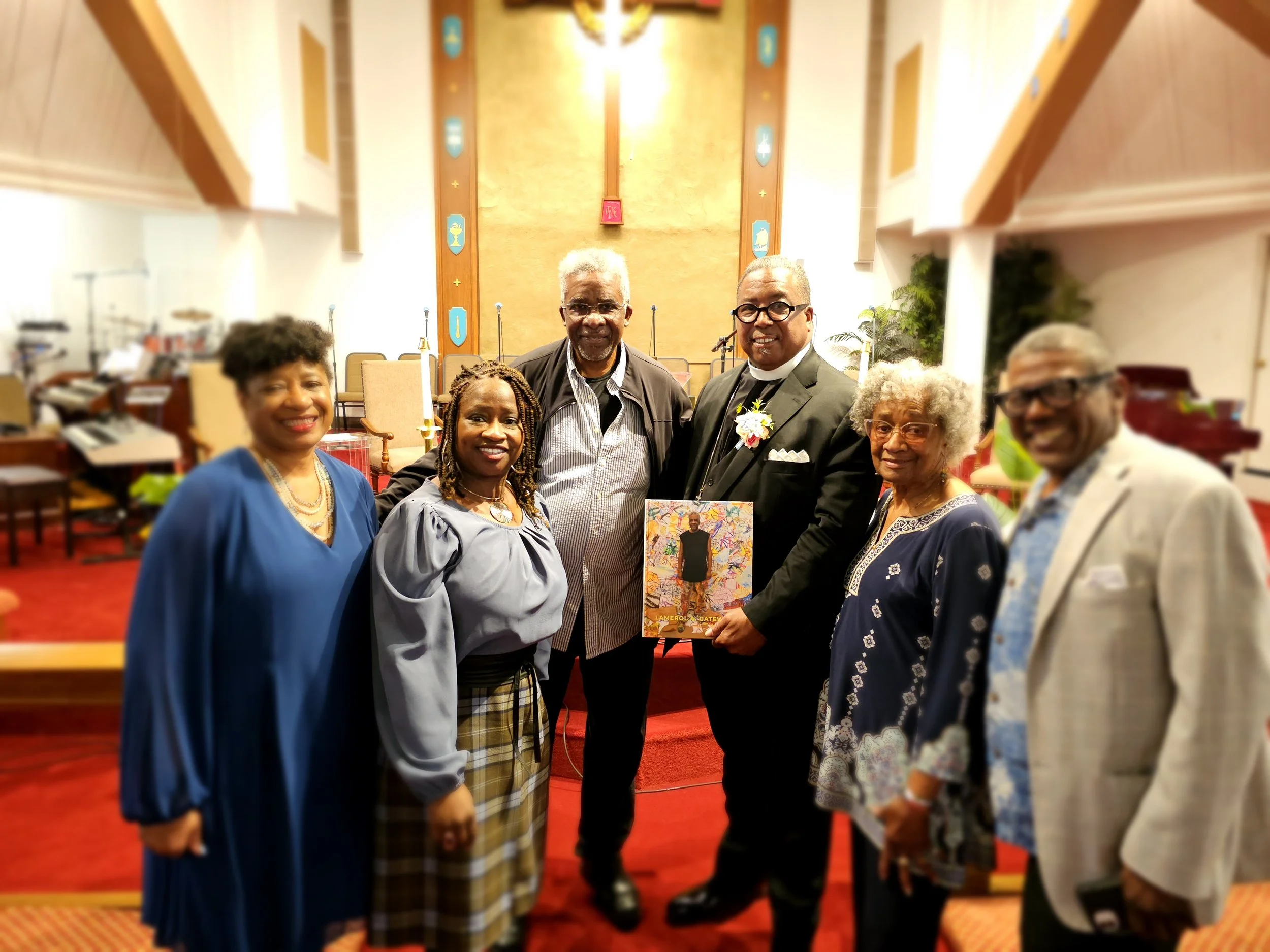 Group of six diverse adults standing inside a church, with an altar and a clock behind them. They are smiling, dressed in semi-formal and formal attire, and appear to be celebrating or attending a special event.