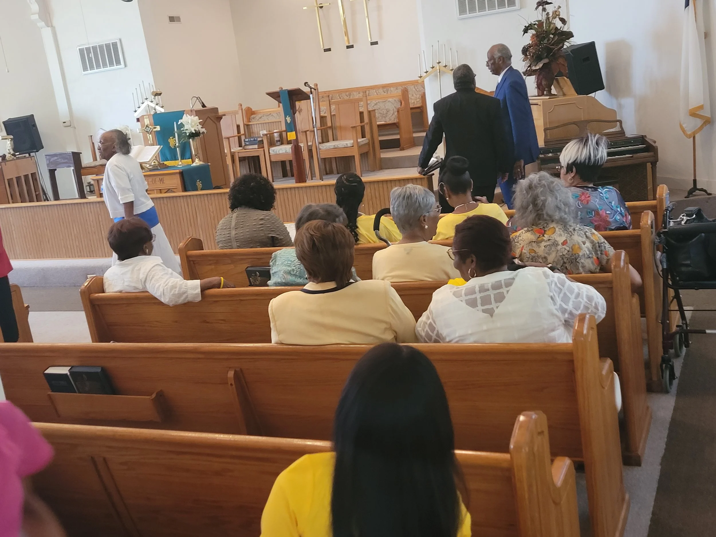 People gathered in a church sanctuary, some sitting on wooden pews and others standing or walking, with an altar and musical instruments in the background.