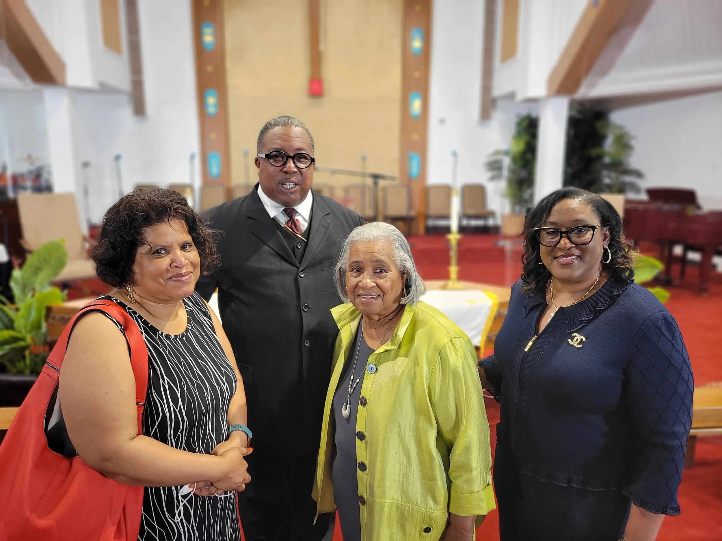Four adults standing inside a church with an altar and a large wooden cross in the background. The group consists of three women and one man, smiling and dressed in formal attire.