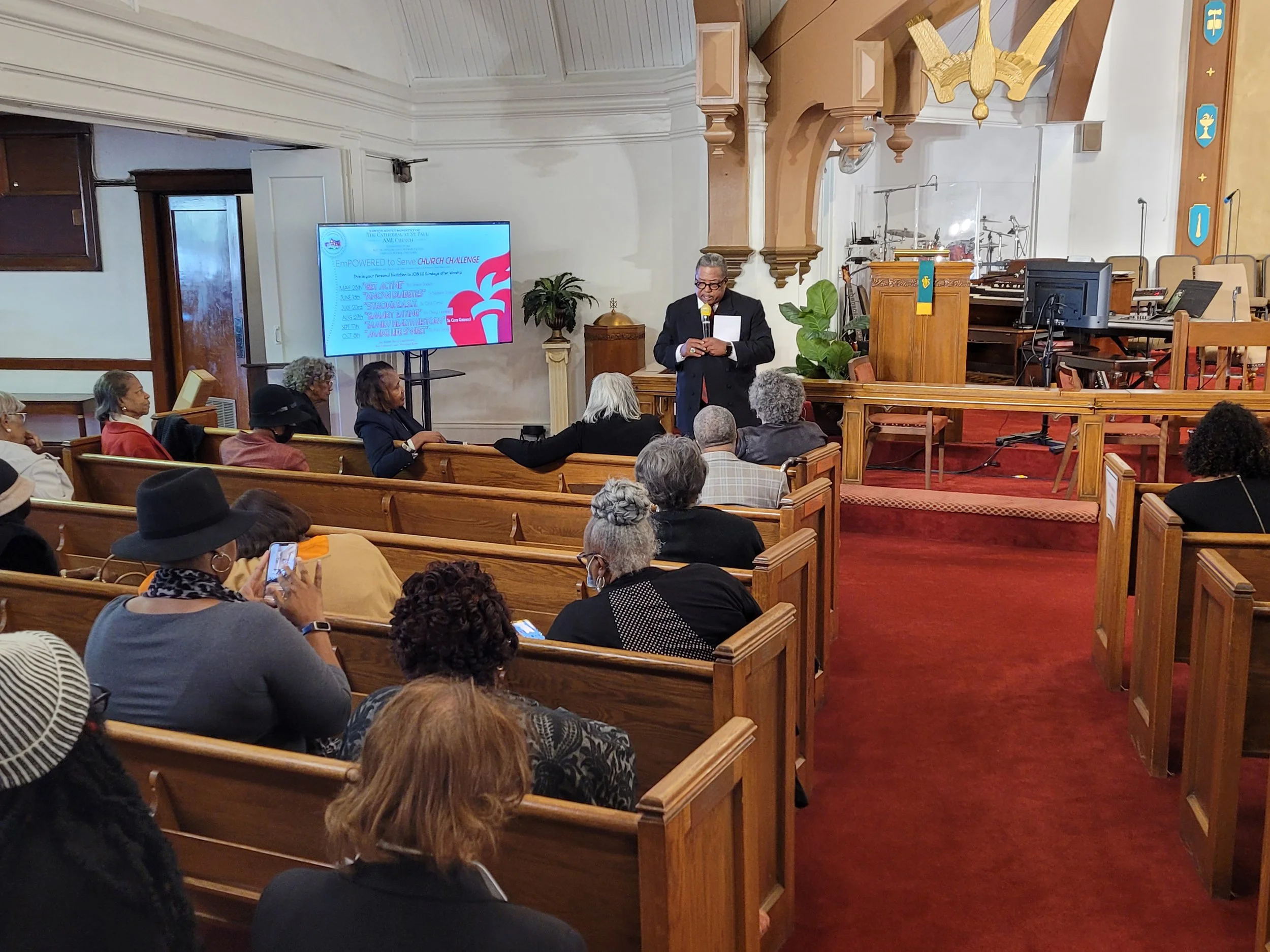A church congregation attending a presentation or lecture inside a church sanctuary with wooden pews, a stage with a speaker, and a large screen displaying presentation slides.