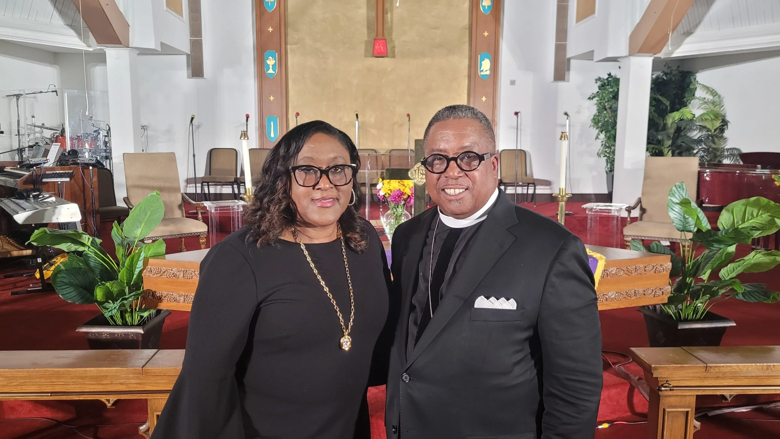 A woman and a man dressed in formal attire posing inside a church with religious symbols, chairs, and musical instruments in the background.