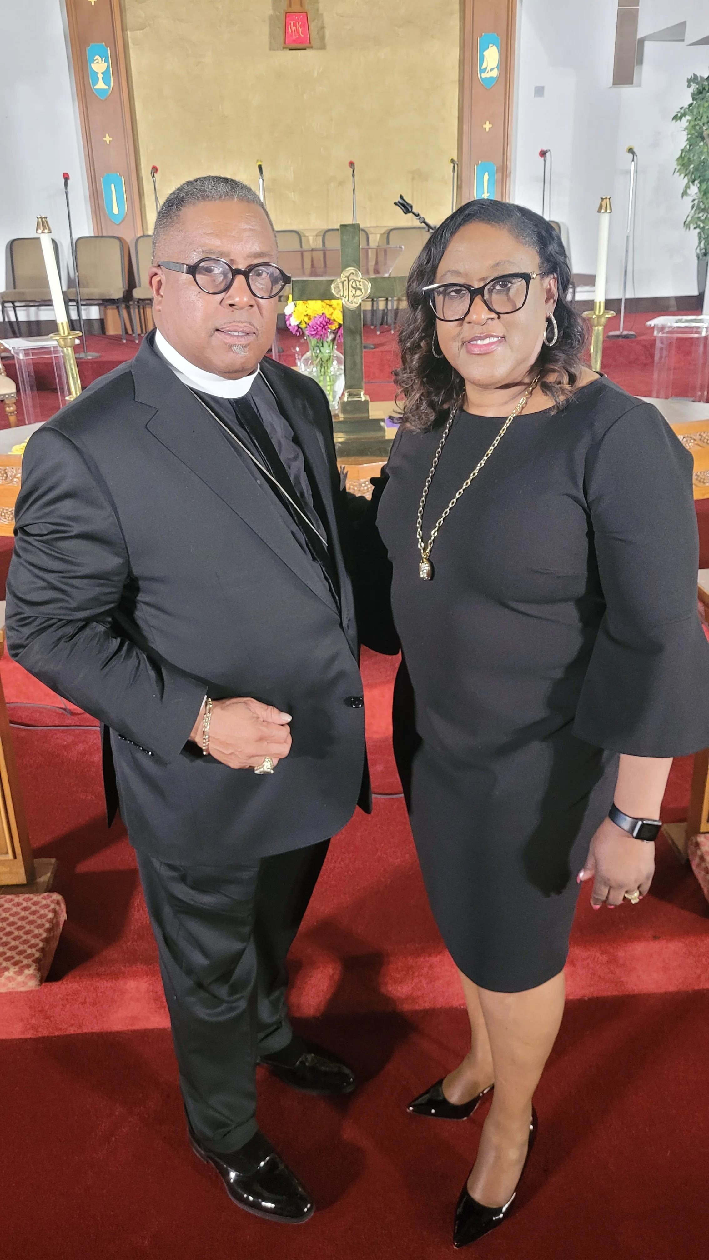 A man and woman standing together inside a church, both dressed in black formal attire. The man is wearing glasses and a clerical collar, while the woman is wearing glasses and a black dress with jewelry. Behind them are church chairs, candlesticks, 