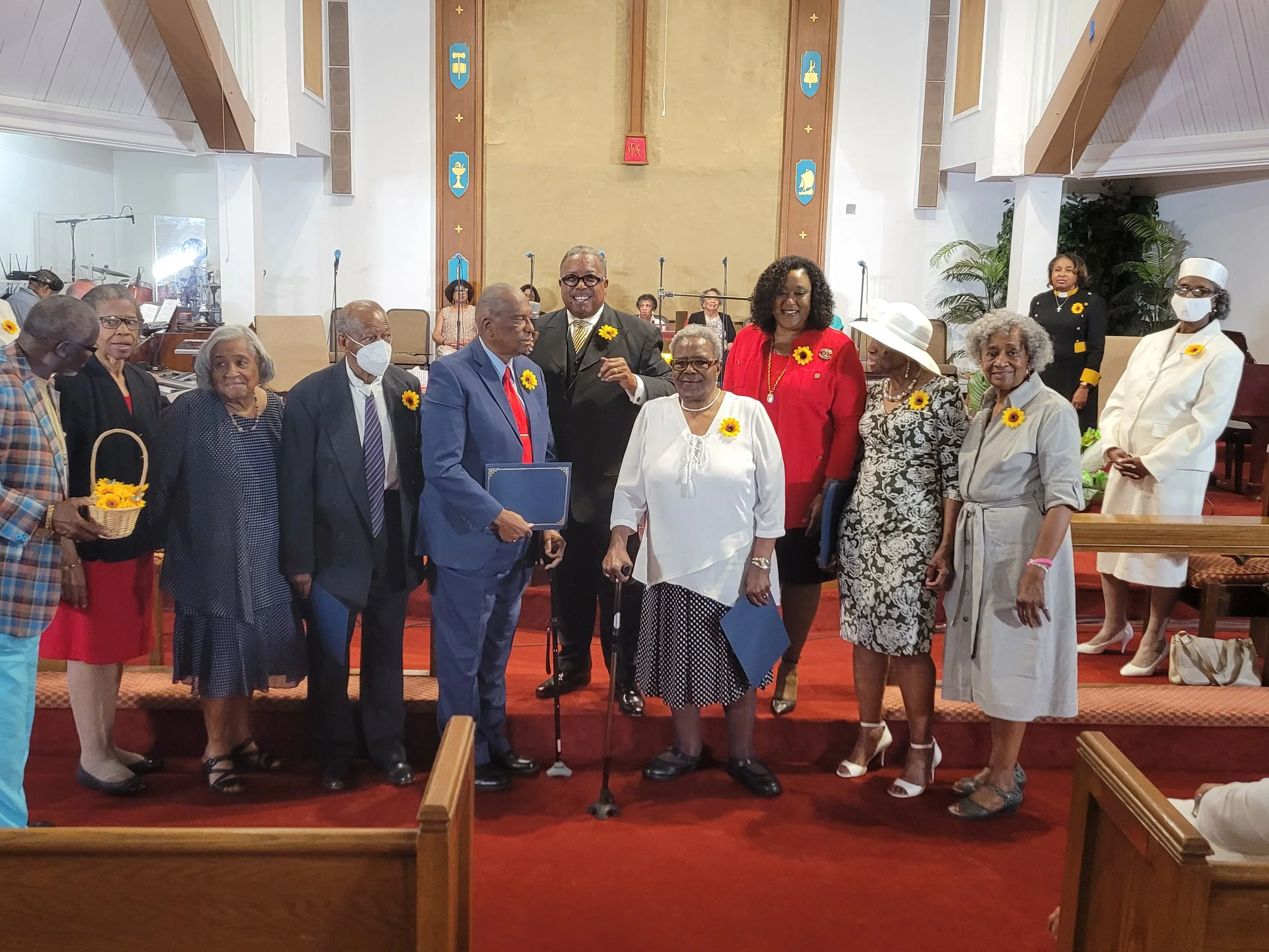 Group of diverse people standing inside a church, some holding certificates or folders, with a man in the center using a cane, all wearing yellow flower pins, during a celebration or recognition event.
