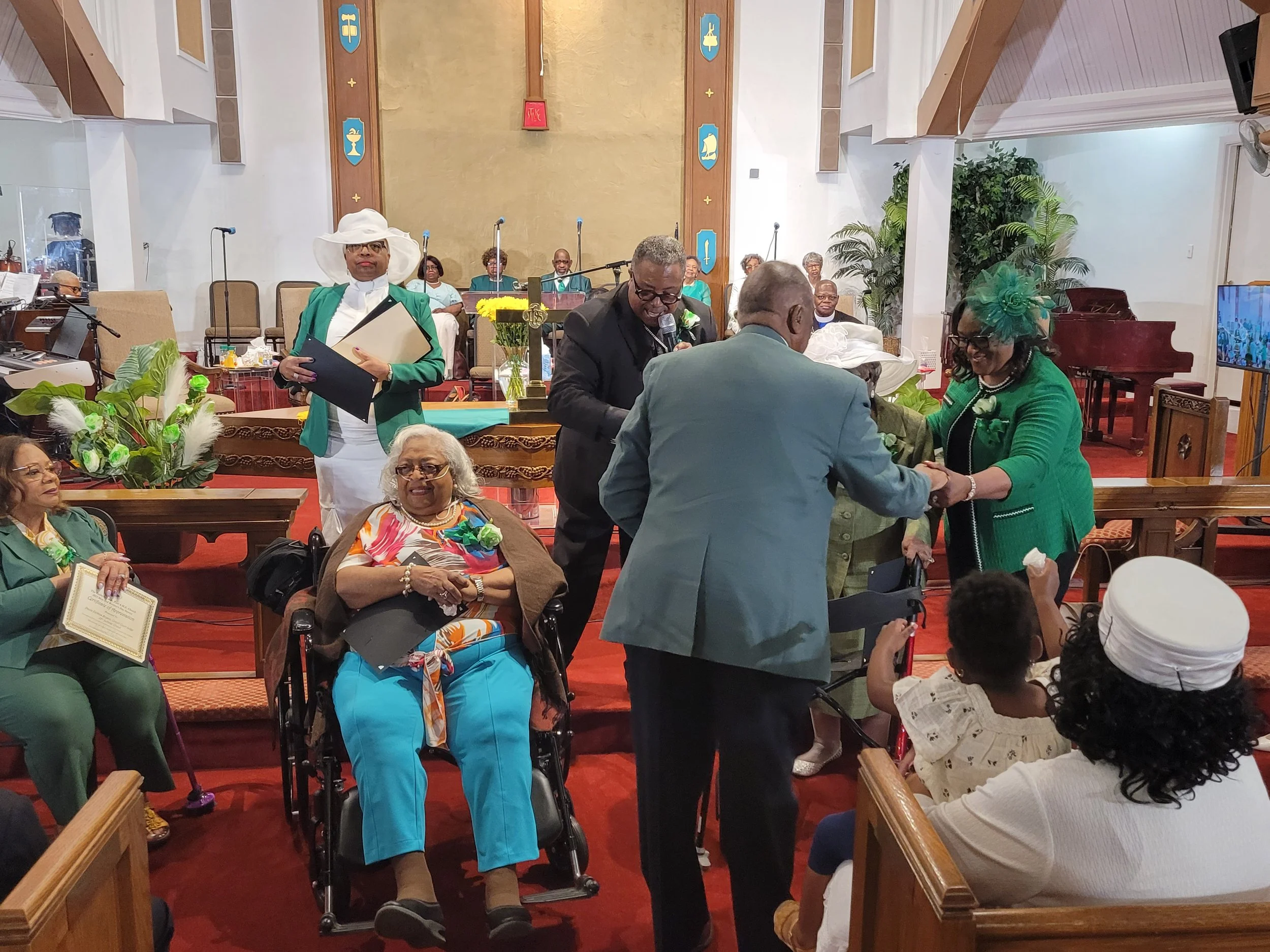 A church service or ceremony taking place, with individuals dressed in formal attire, some wearing hats. People are engaged in handshakes and holding certificates or documents. The decor includes flowers and religious symbols.