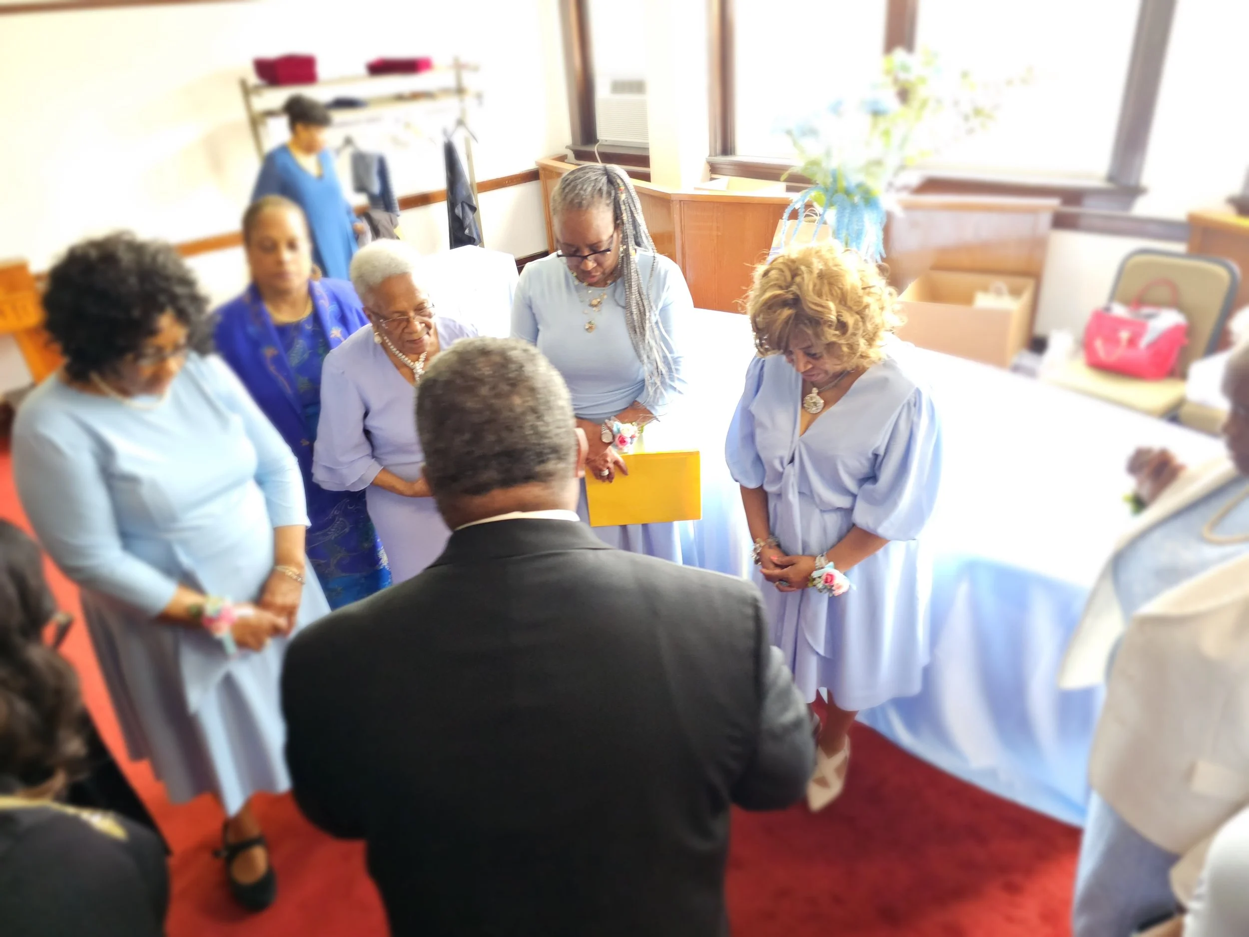Group of seven women and one man in formal attire praying in a room with red carpet, windowed walls, and minimal decor.