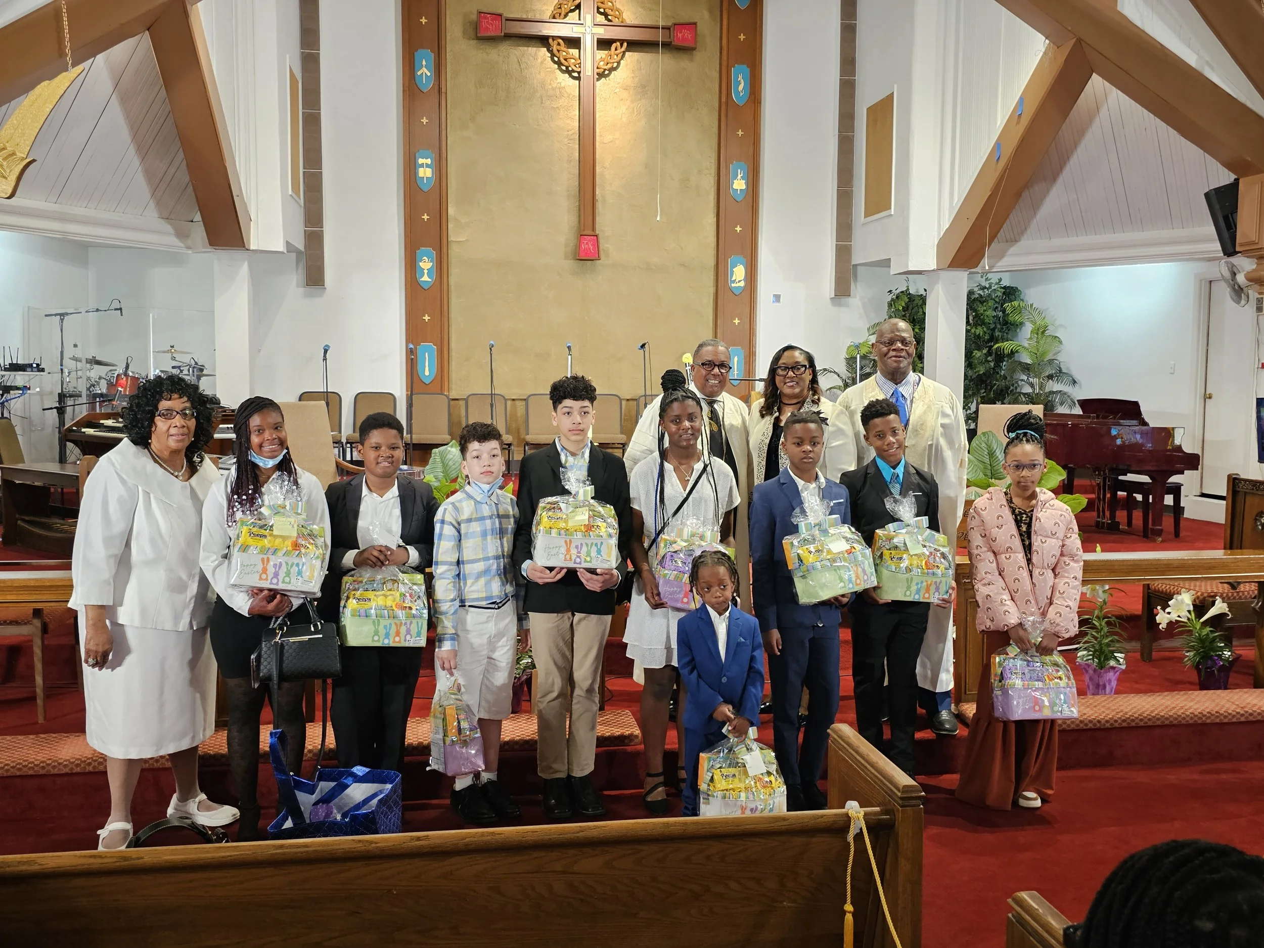 Group of children and adults standing on stage in a church, holding gift baskets, with a cross and religious symbols on the wall behind them.