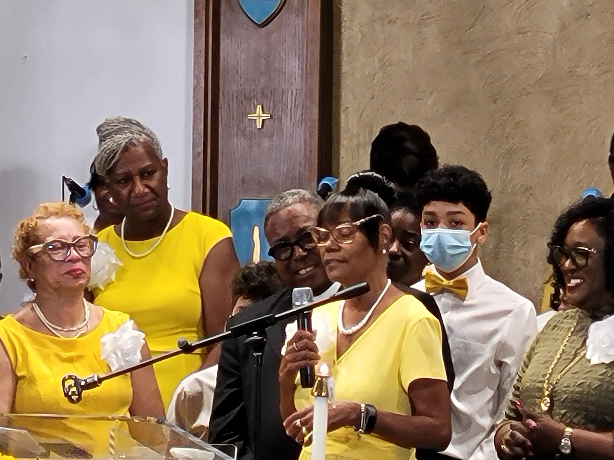 Group of people at an indoor event, some women wearing yellow dresses, a woman speaking into a microphone, a young man wearing a face mask, and others observing, in a room with a textured beige wall.