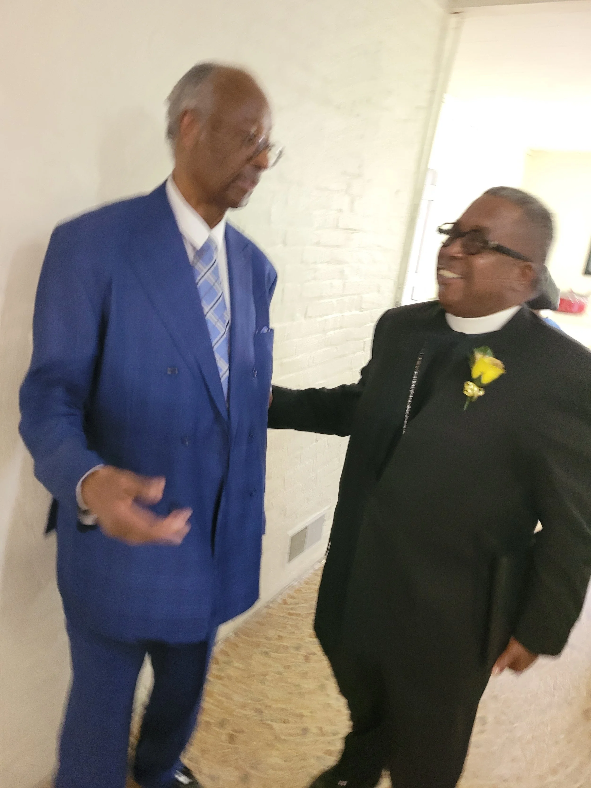 Two men shaking hands and smiling, one wearing a blue suit and the other in a black clergy outfit with a yellow boutonniere, inside a building with a white brick wall.