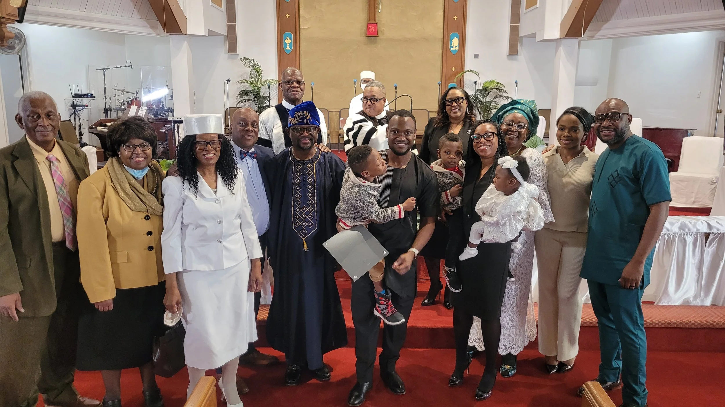 A group photo of people in a church with some children, all dressed in formal and cultural attire, standing on a red carpeted platform.