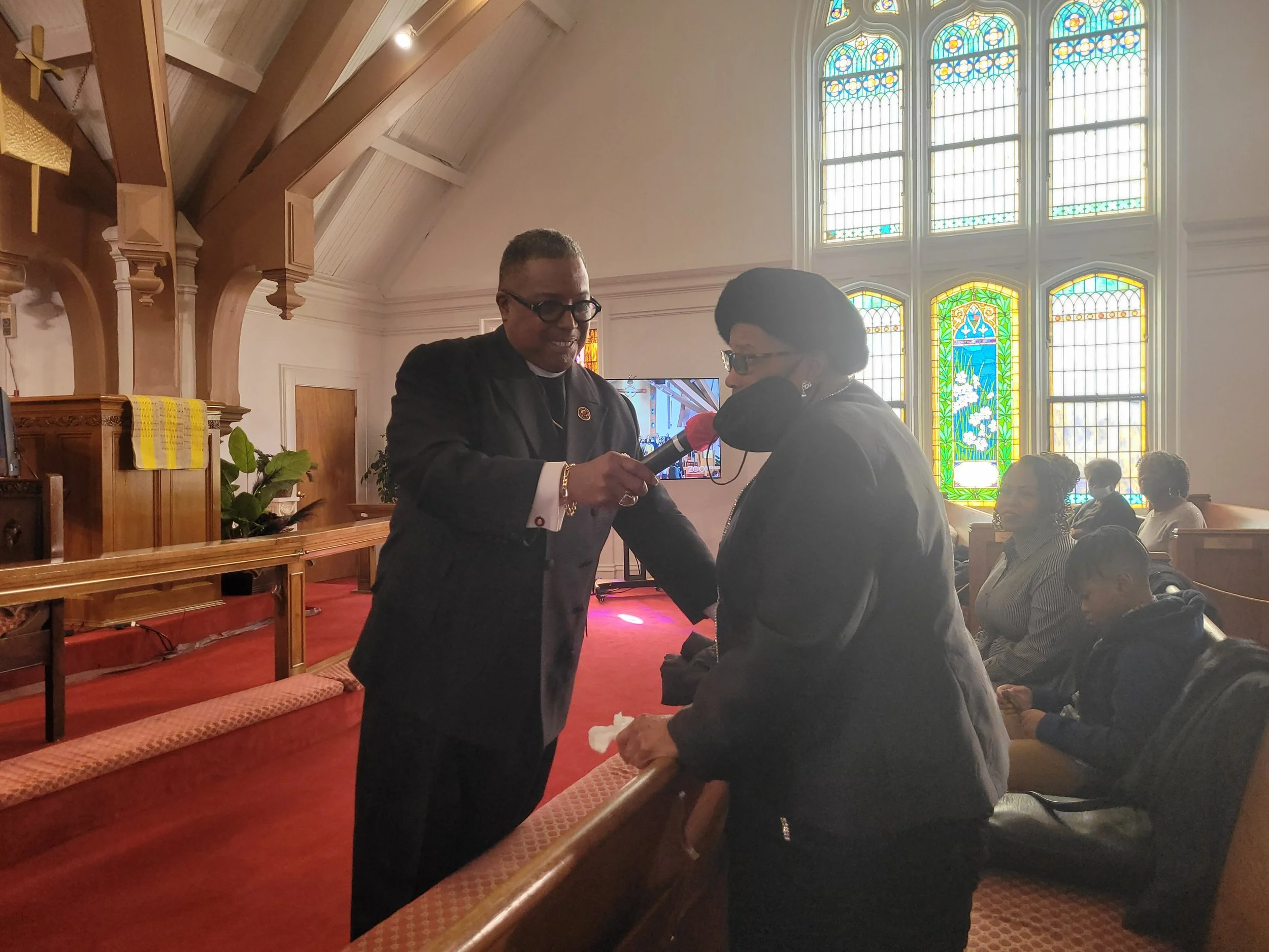 A woman at an outdoor church service being interviewed by a man holding a microphone, with stained glass windows in the background.