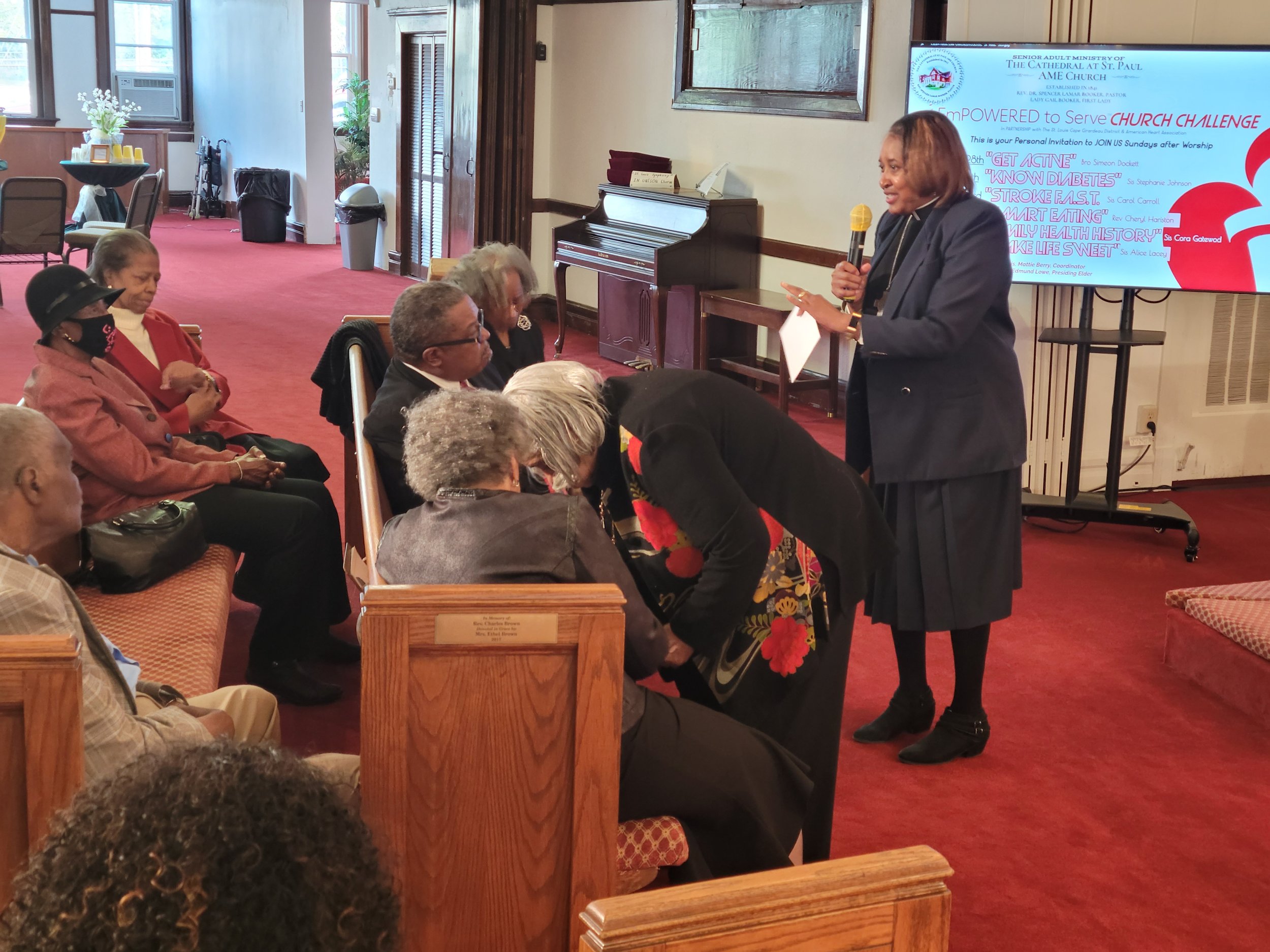 A woman delivering a speech with a microphone to an audience of older adults seated in a room, with a presentation screen displaying information related to a church challenge.