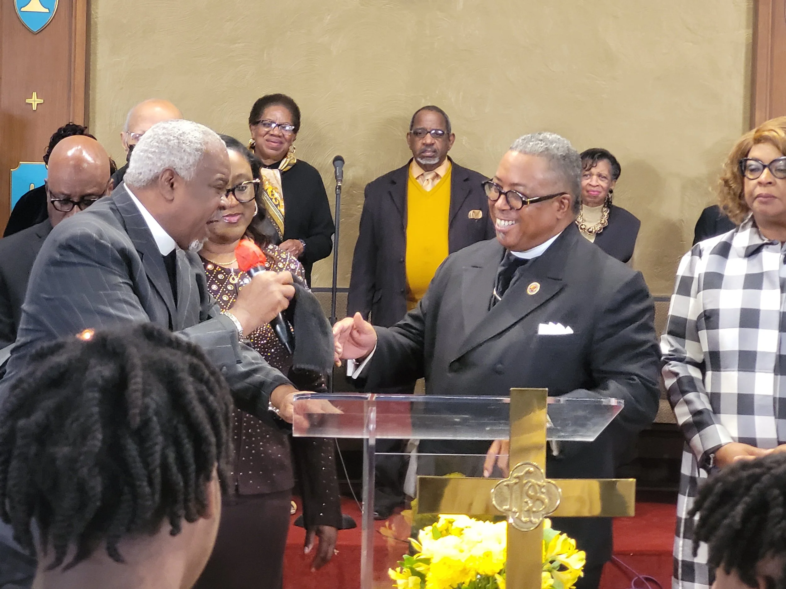 A group of people at a church service or ceremony, with two men shaking hands at the altar. One man is older with gray hair, wearing a gray suit, and the other is younger with gray hair and glasses, dressed in a black suit with a clerical collar. The