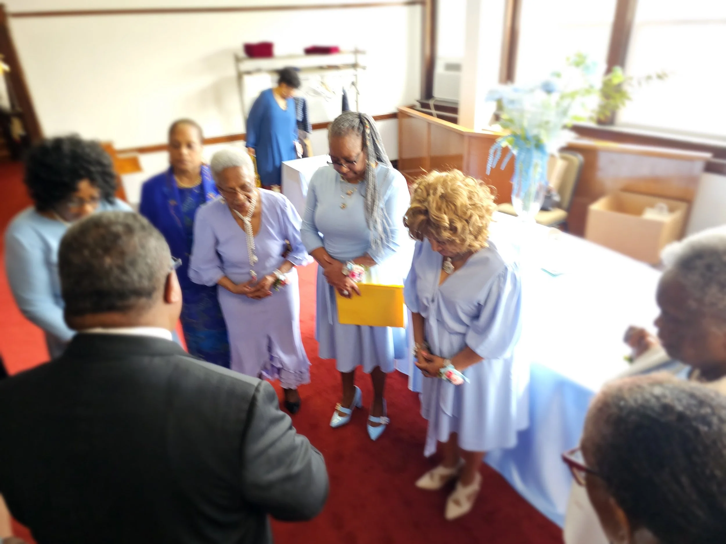 Group of women and men standing in a circle in a room, with some looking downward and others looking at a man in a suit, in a gathering or ceremony setting.