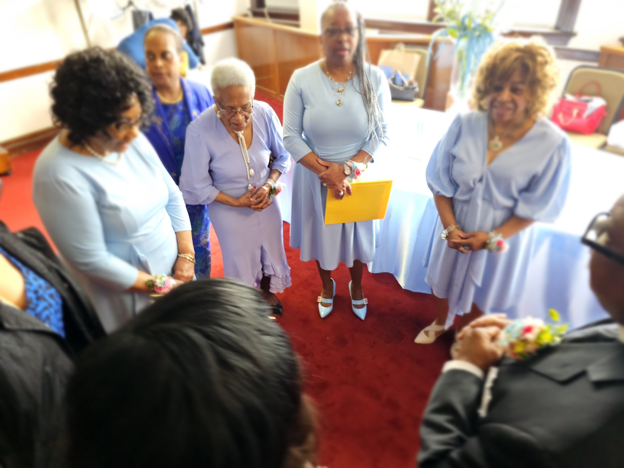 Group of women standing together in a circle, dressed in light blue clothing, inside a room with a red carpet, engaged in a prayer or moment of reflection.