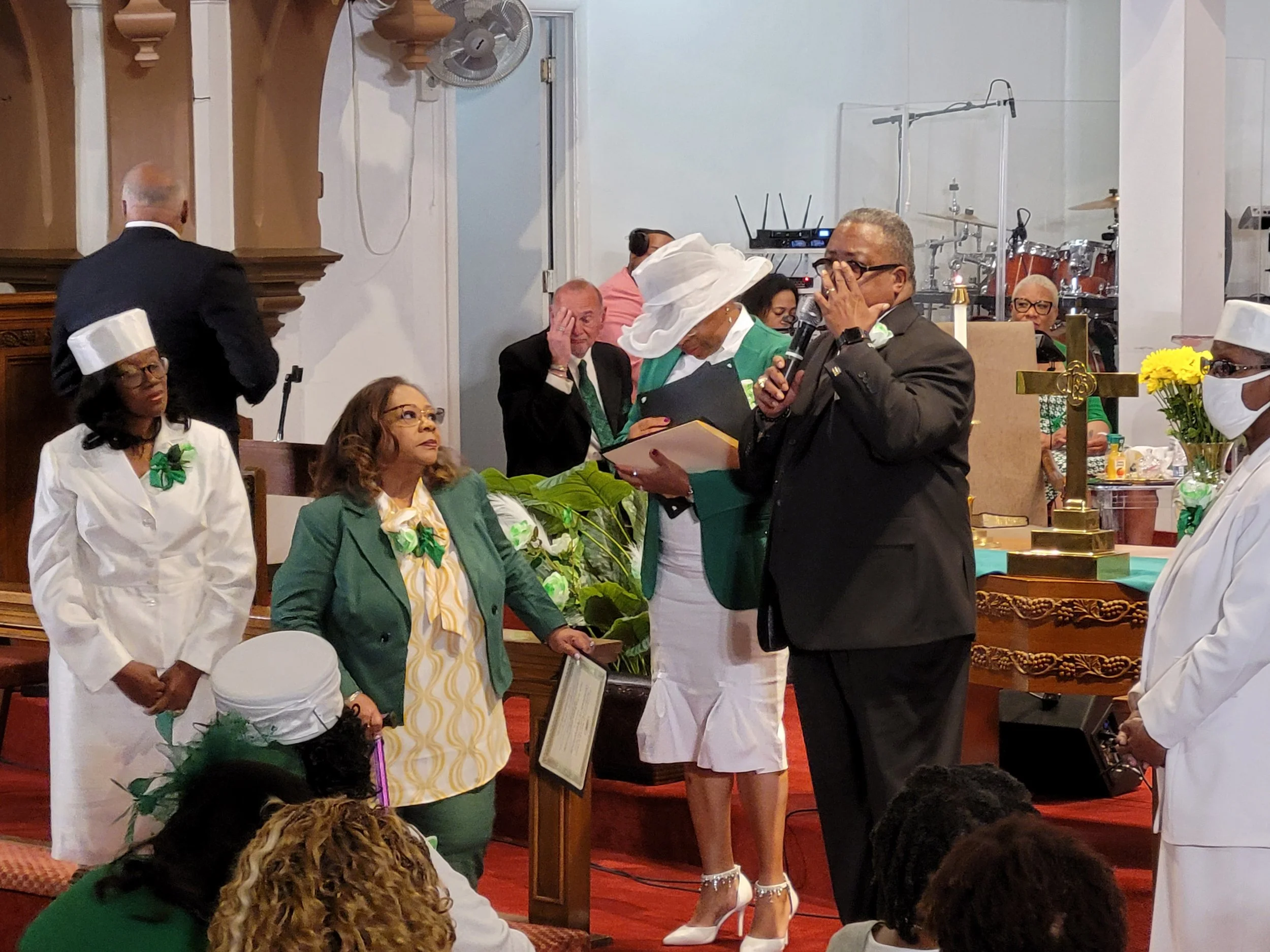 A religious ceremony or service taking place in a church, with several clergy members and officials present. Women are dressed in white and green, some with hats and masks, and a man is speaking into a microphone while holding a book or document.