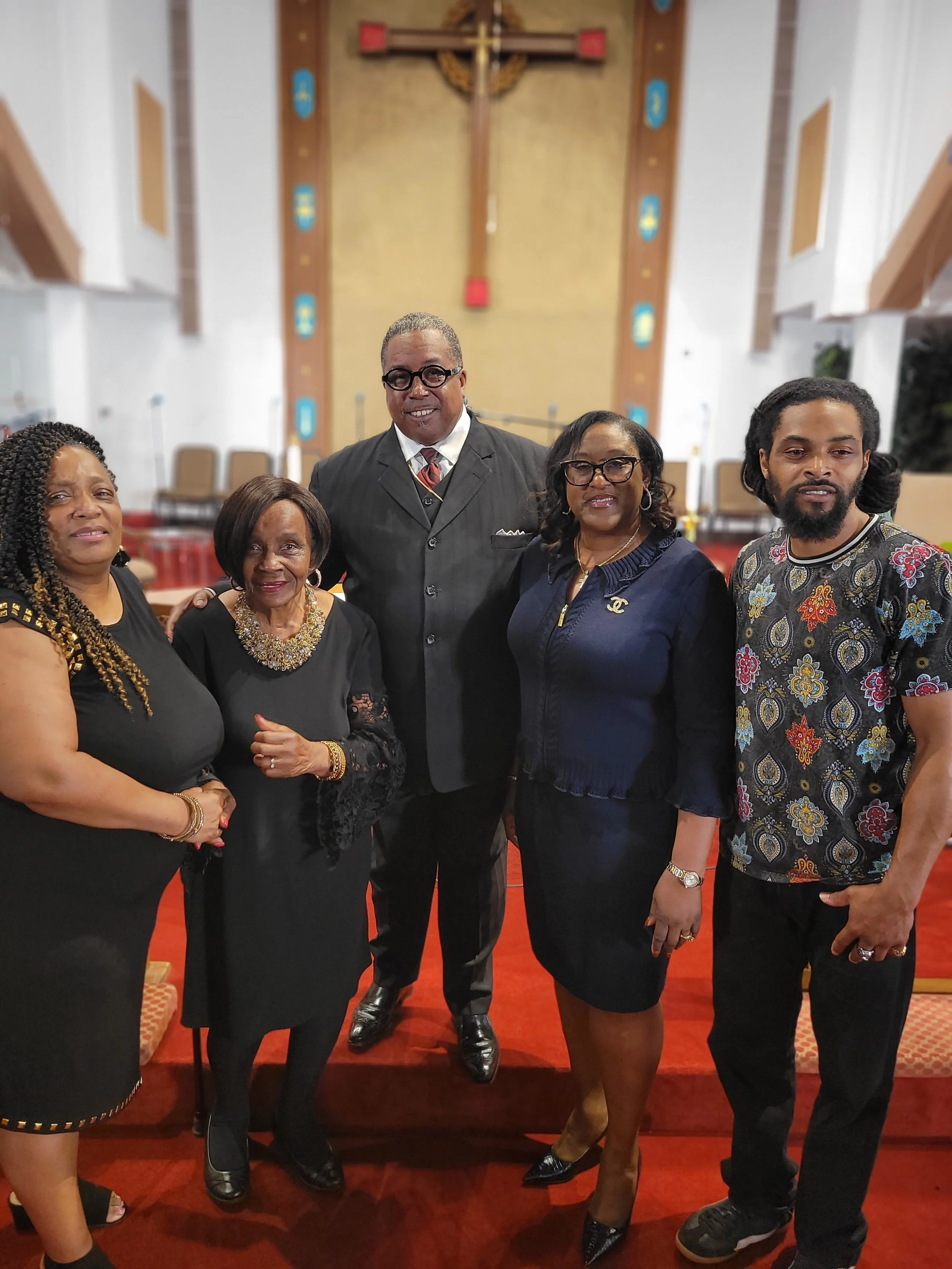 Group of six people standing inside a church, with a cross in the background and red carpet on the floor.
