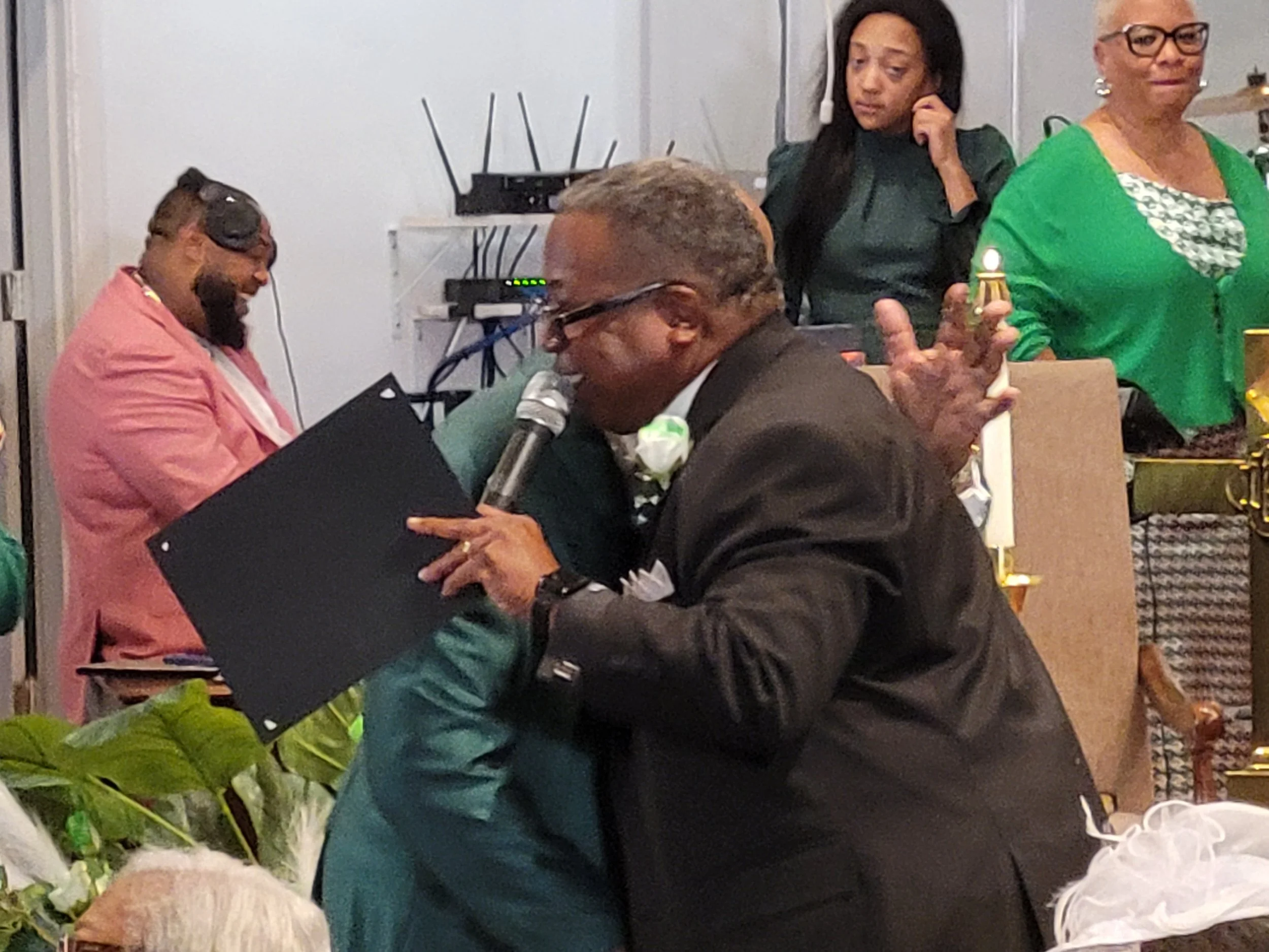 A man and a woman are hugging at a celebration, with people in the background, some smiling and others watching. The setting appears to be a festive indoor event.