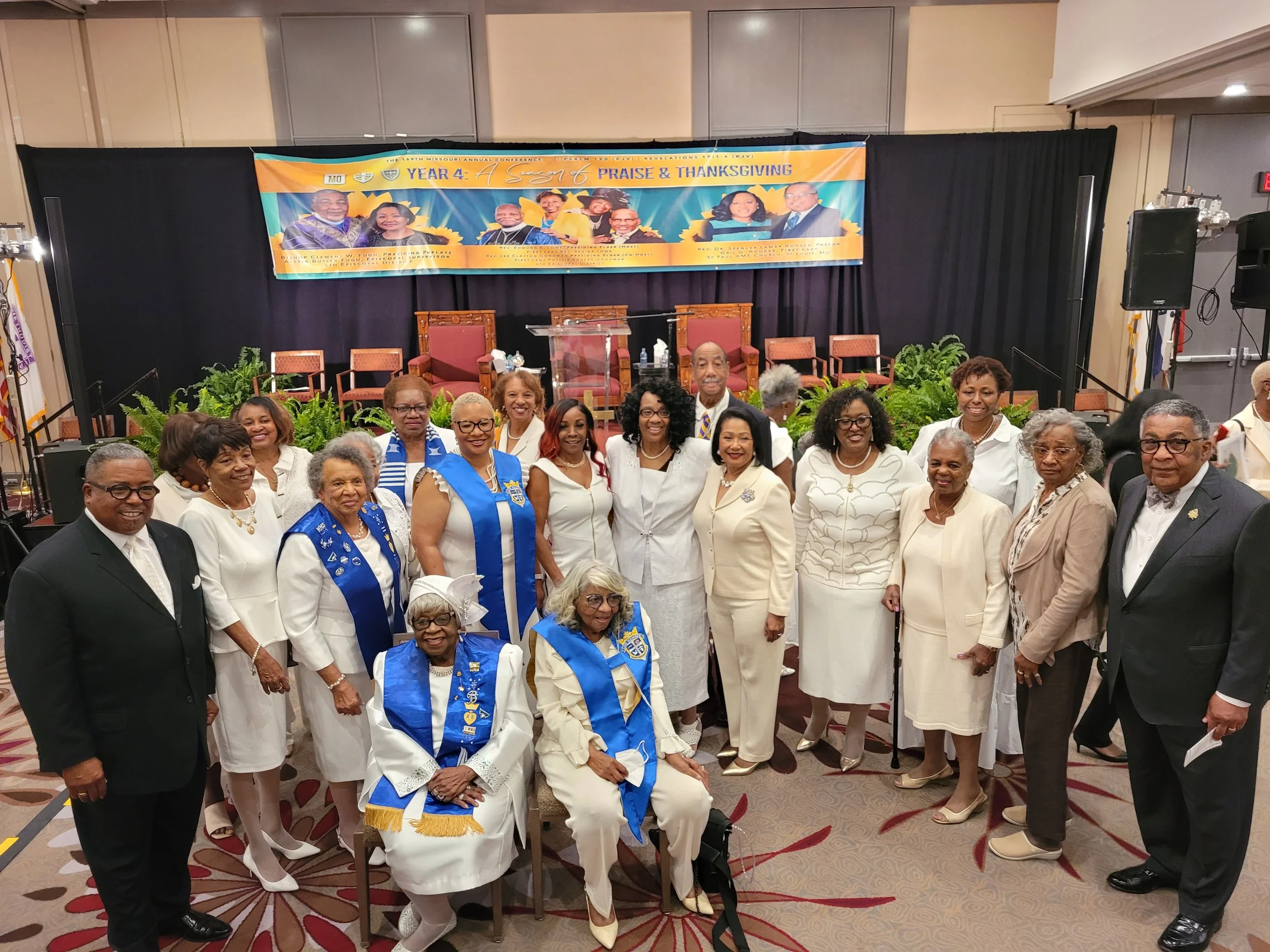 Group photo of diverse men and women in formal attire, some wearing blue sashes or vests with insignia, standing on a stage with a banner behind them that reads 'Praise & Thanksgiving' and features images of people. The setting appears to be an event